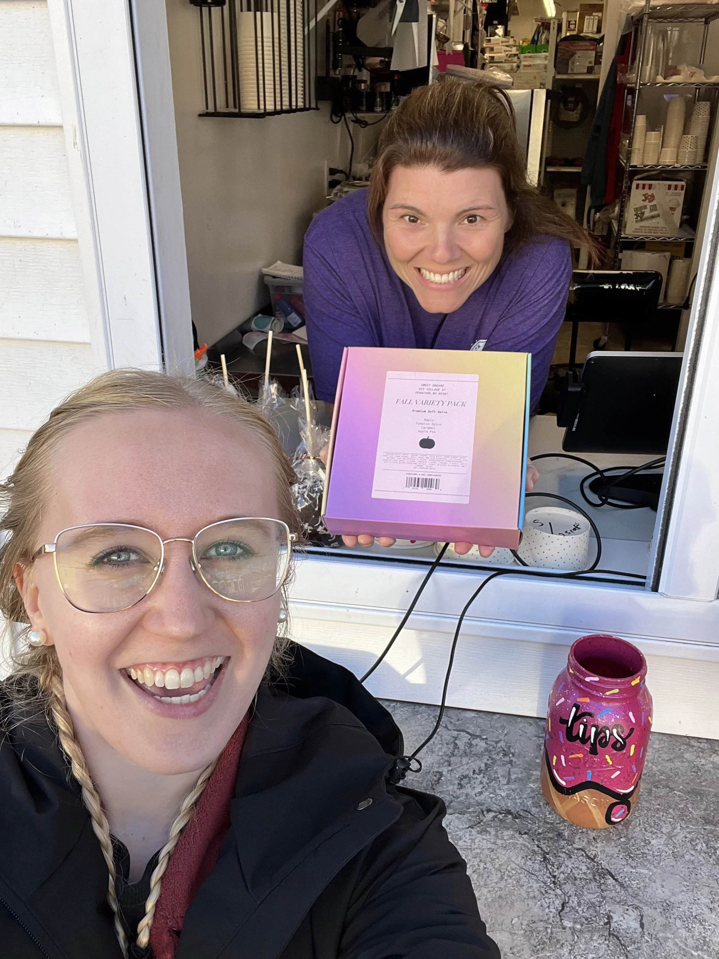 Two women smiling at a food stand window. The woman in the foreground, wearing glasses and a black jacket, has blond hair styled in a braid. The woman behind the window, wearing a purple shirt, has brown hair and is holding a colorful box labeled 'Fall Variety Pack'. A pink jar with a lid labeled 'Tips' is on the counter.