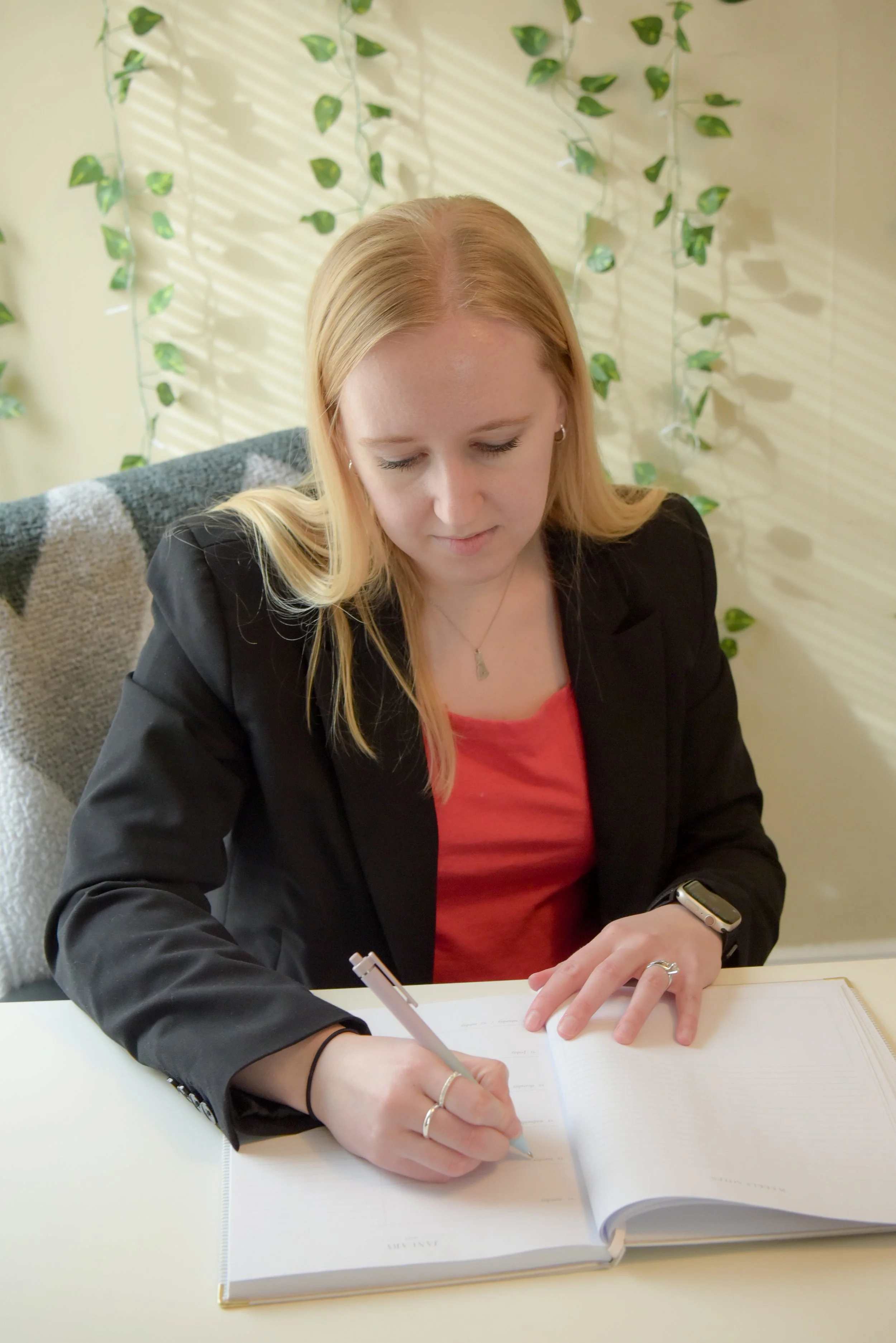 A woman with long blonde hair, wearing a black blazer and red top, sitting at a table, writing in a notebook with a pen, with greenery and shadow patterns on the wall in the background.