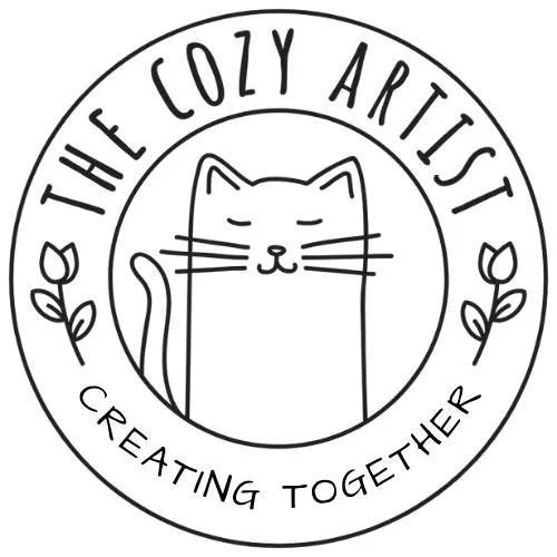Logo featuring a simple line drawing of a smiling cat inside a circle, with the text 'The Cozy Artist' above and 'Creating Together' below, and decorative leaves on either side.