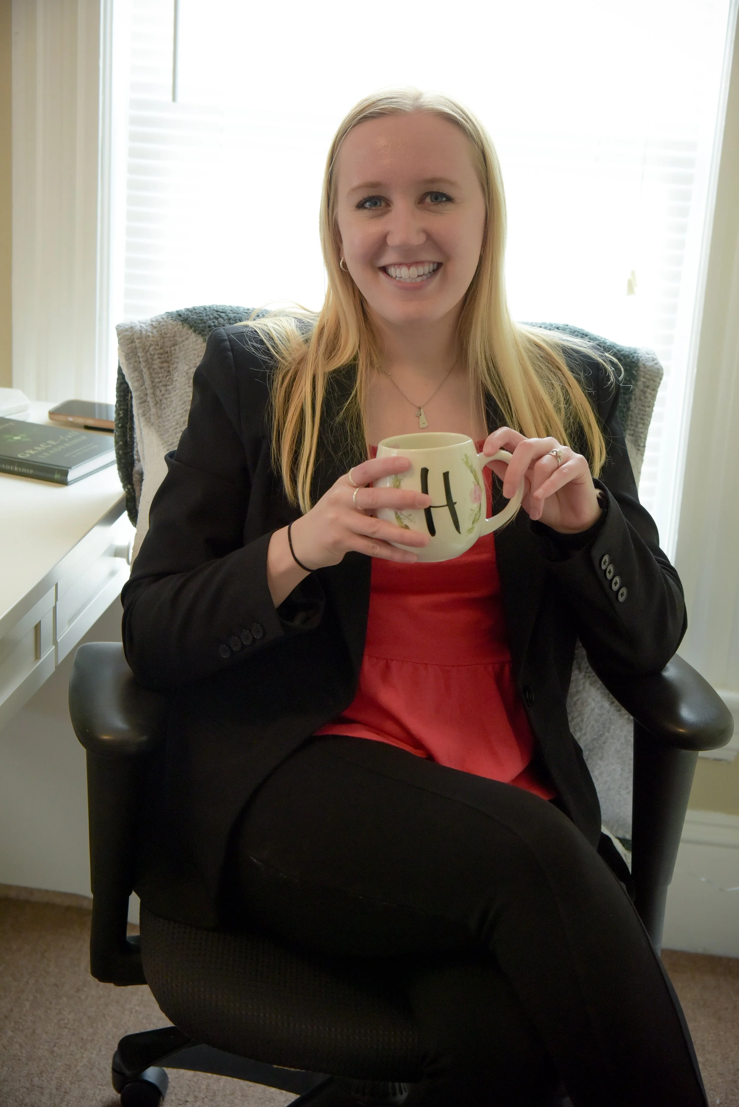 A woman with long blonde hair smiling while sitting in a black office chair, holding a white mug with a letter 'H' on it, in an office with a window and a desk with books in the background.