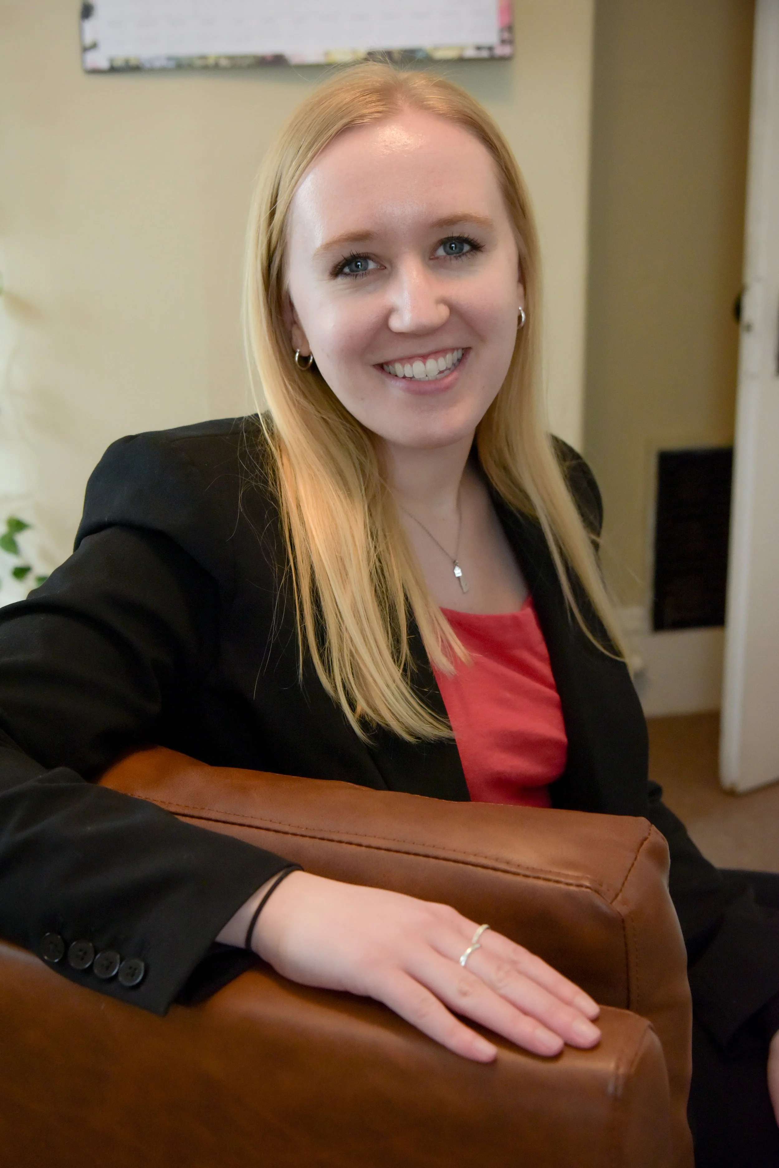 A young woman with blonde hair, blue eyes, and earrings, smiling for a selfie, sitting at a table with her hand resting on a brown purse. She is wearing a black blazer over a red top.