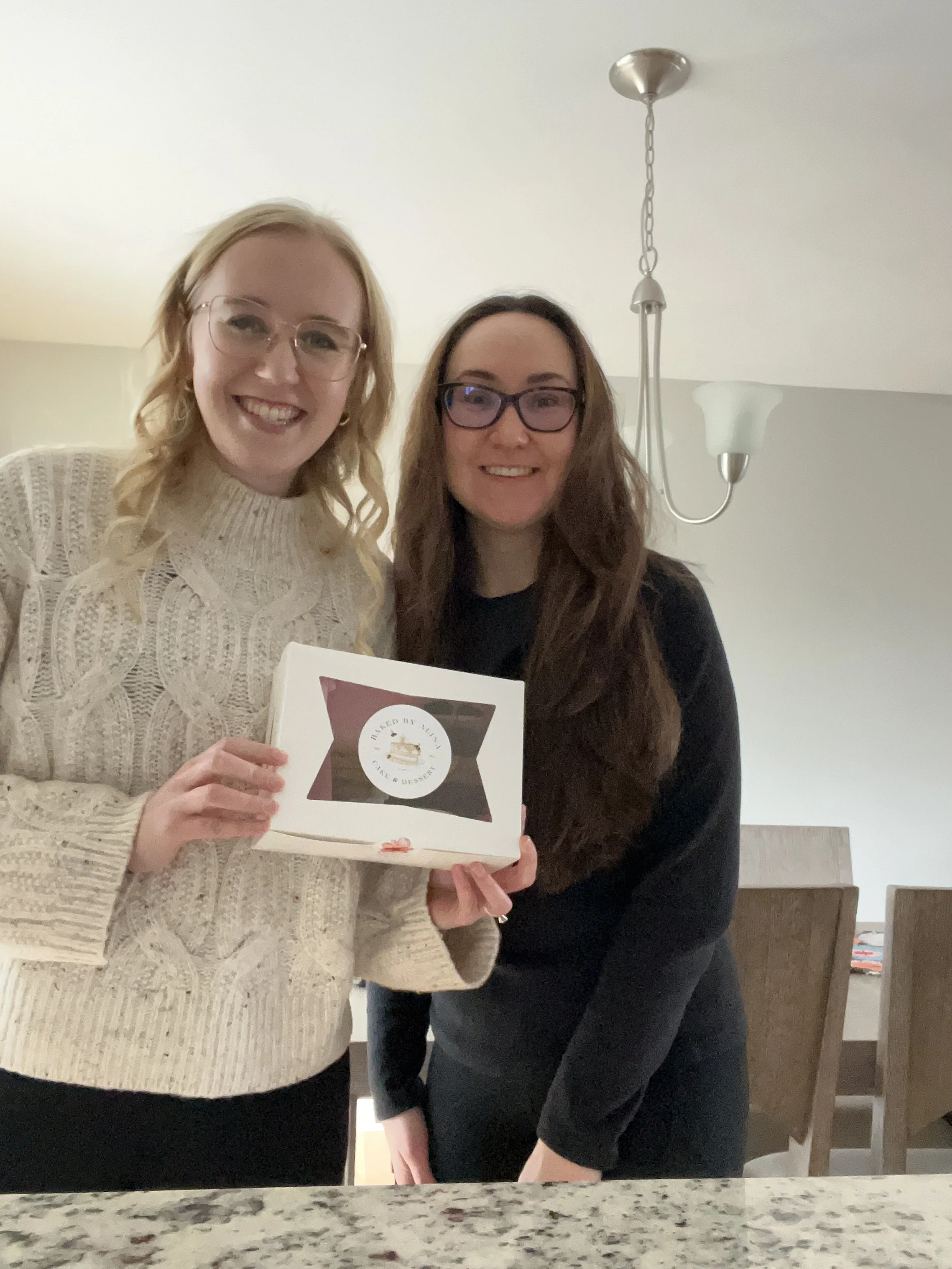 Two women smiling and holding a box labeled 'Baked by Yuki Cake & Dessert' in a kitchen.