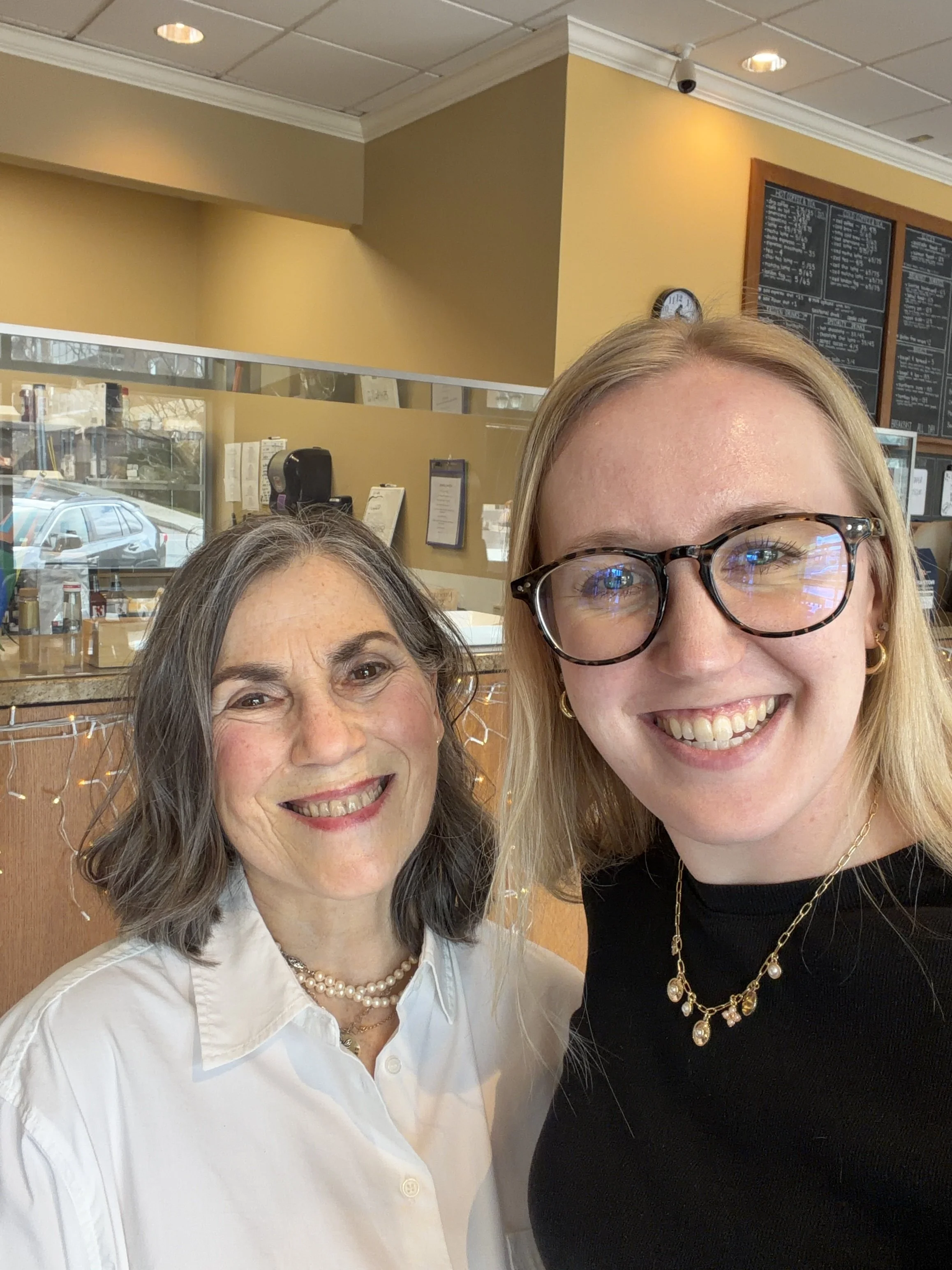 Two women smiling for a selfie inside a cafe. One woman has curly gray hair wearing a white blouse and pearls, the other has straight blonde hair wearing glasses, a black top, and gold jewelry. A menu board and a clock are visible in the background.
