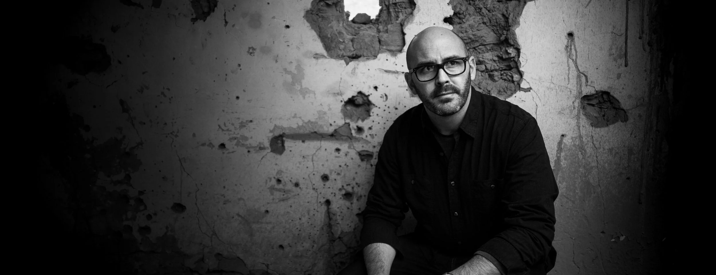 A black-and-white photo of a man with glasses, a beard, and a bald head sitting against a distressed wall with holes and cracks.