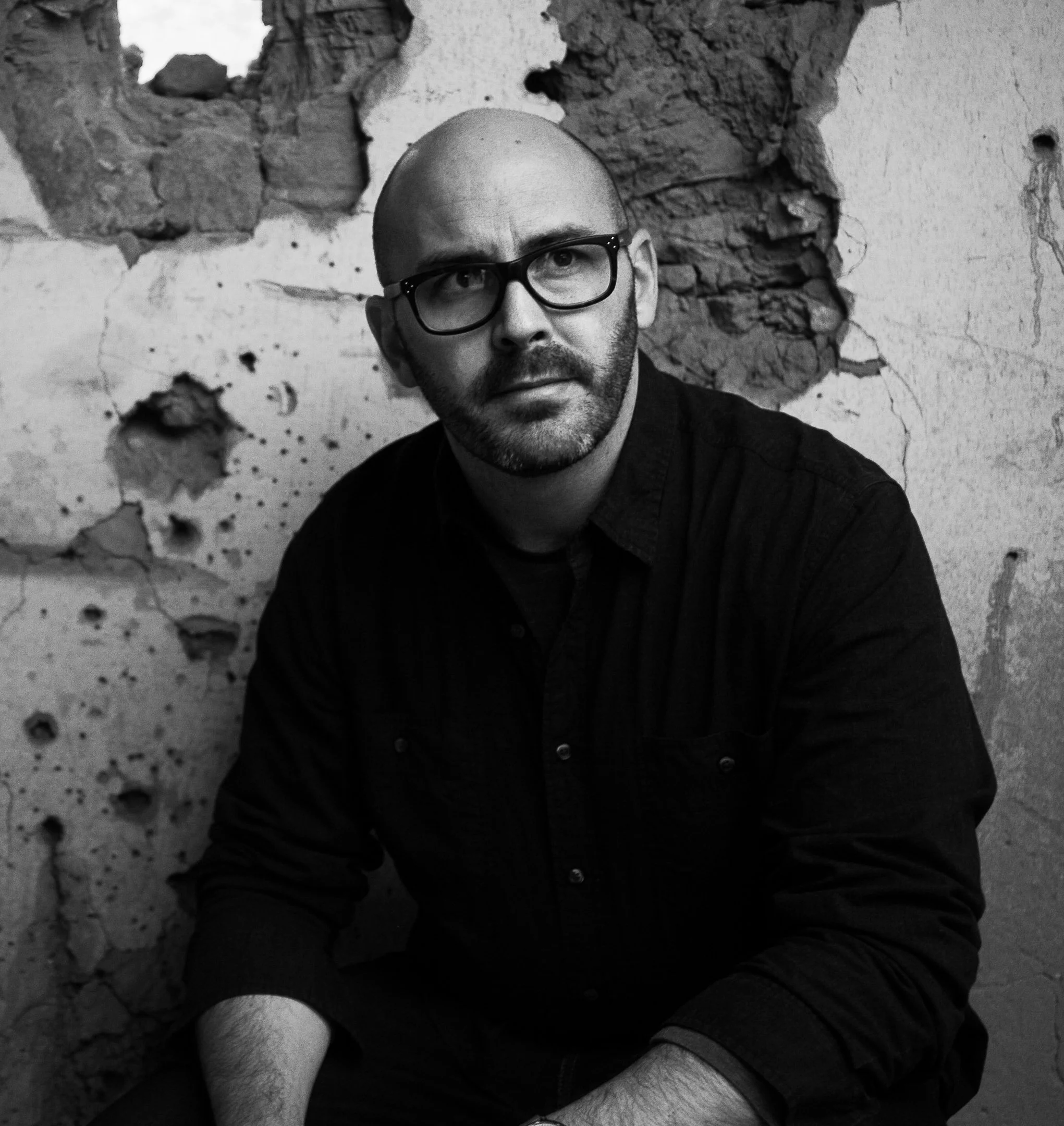 A black and white photo of a man, director, with glasses and a beard, sitting on set with a weathered, broken wall with exposed bricks behind him.