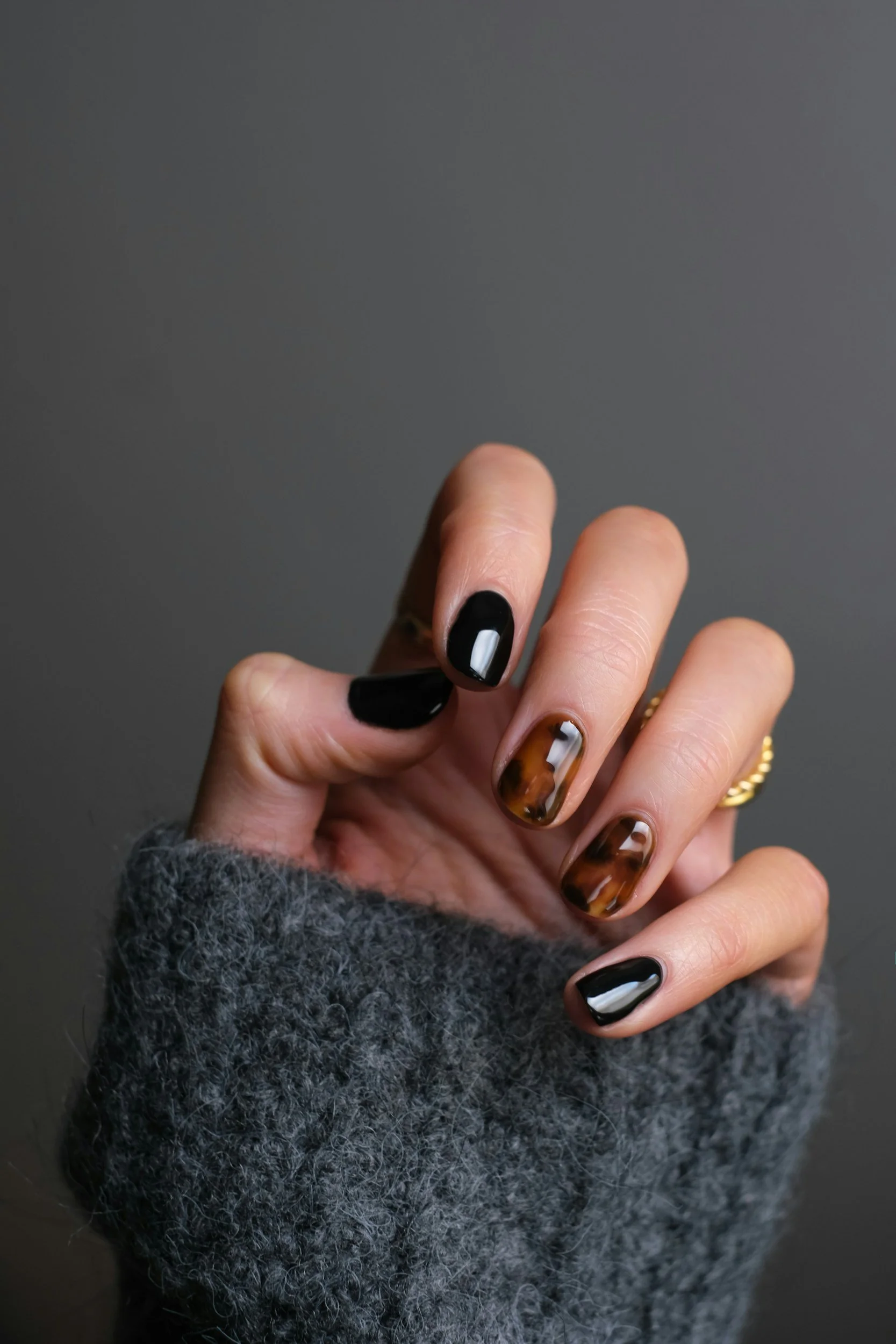 Close-up of a hand with manicured nails, some painted black and others with tortoiseshell design, against a plain gray background, wearing a gray fuzzy sweater.