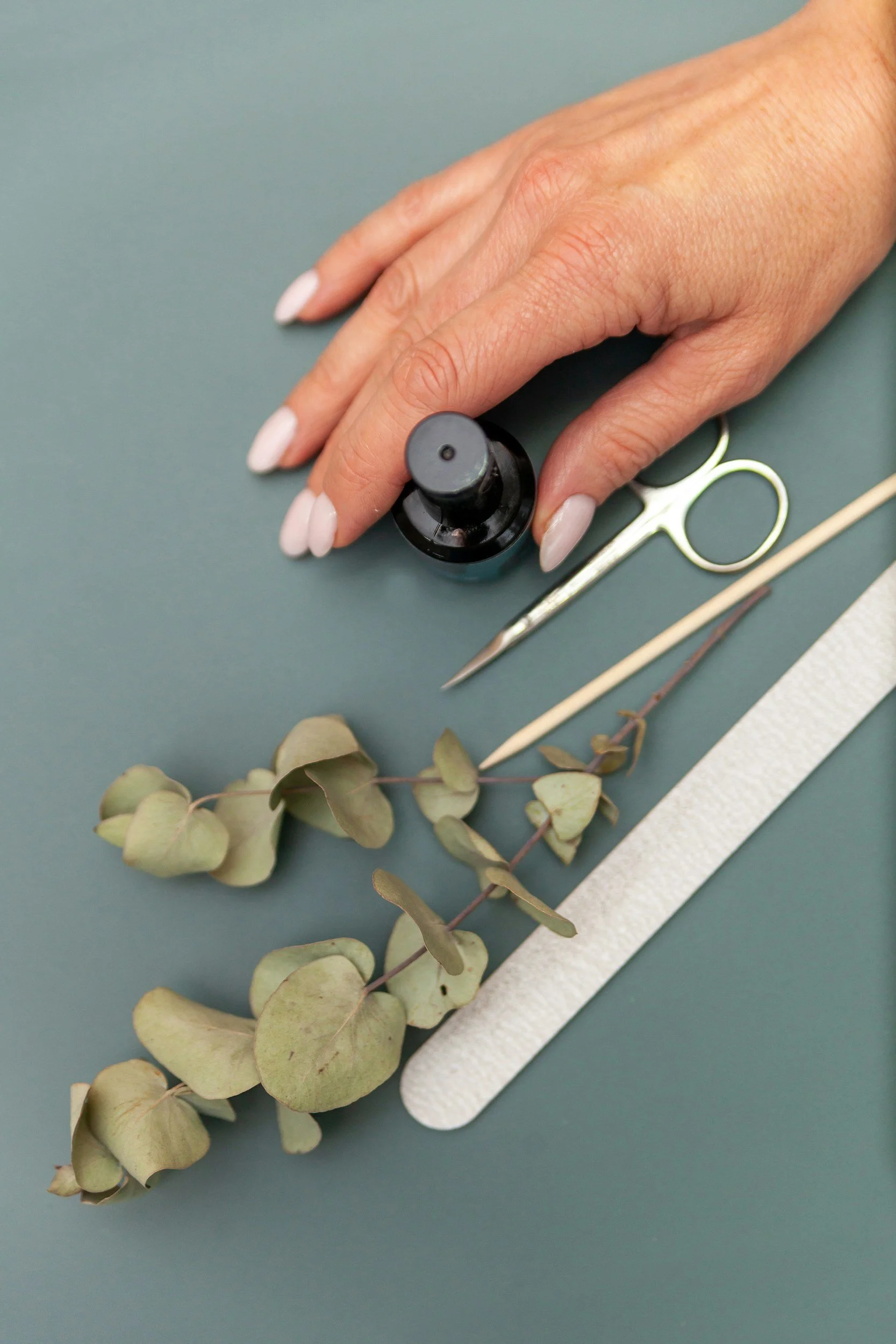 A hand holding a small black essential oil bottle on a green surface, with manicure, scissors, tweezers, and eucalyptus leaves nearby.