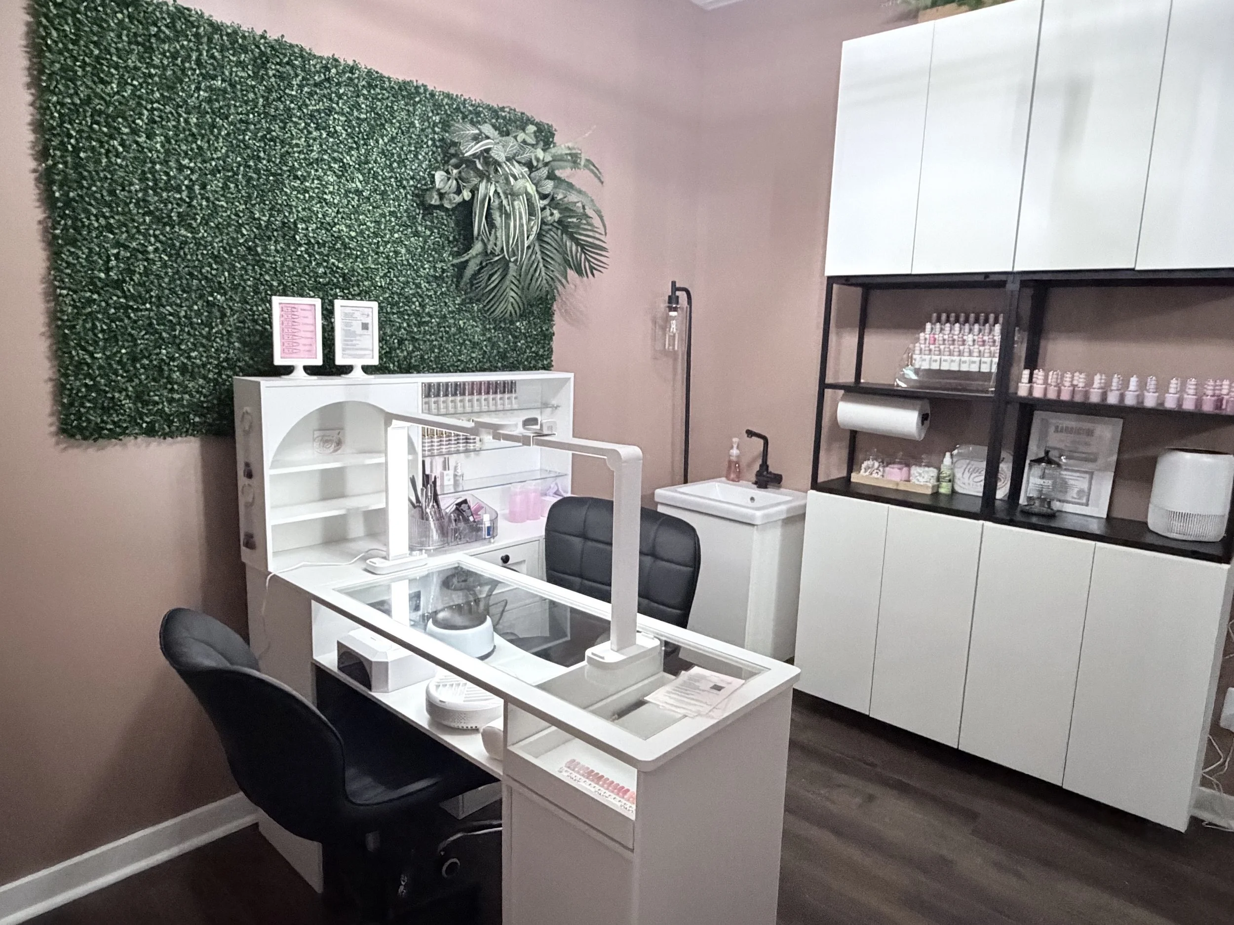 A nail salon workspace with a white manicure station, black swivel chair, and various nail and beauty products on black shelving units. A wall decorated with green artificial plants and a pink wall background.
