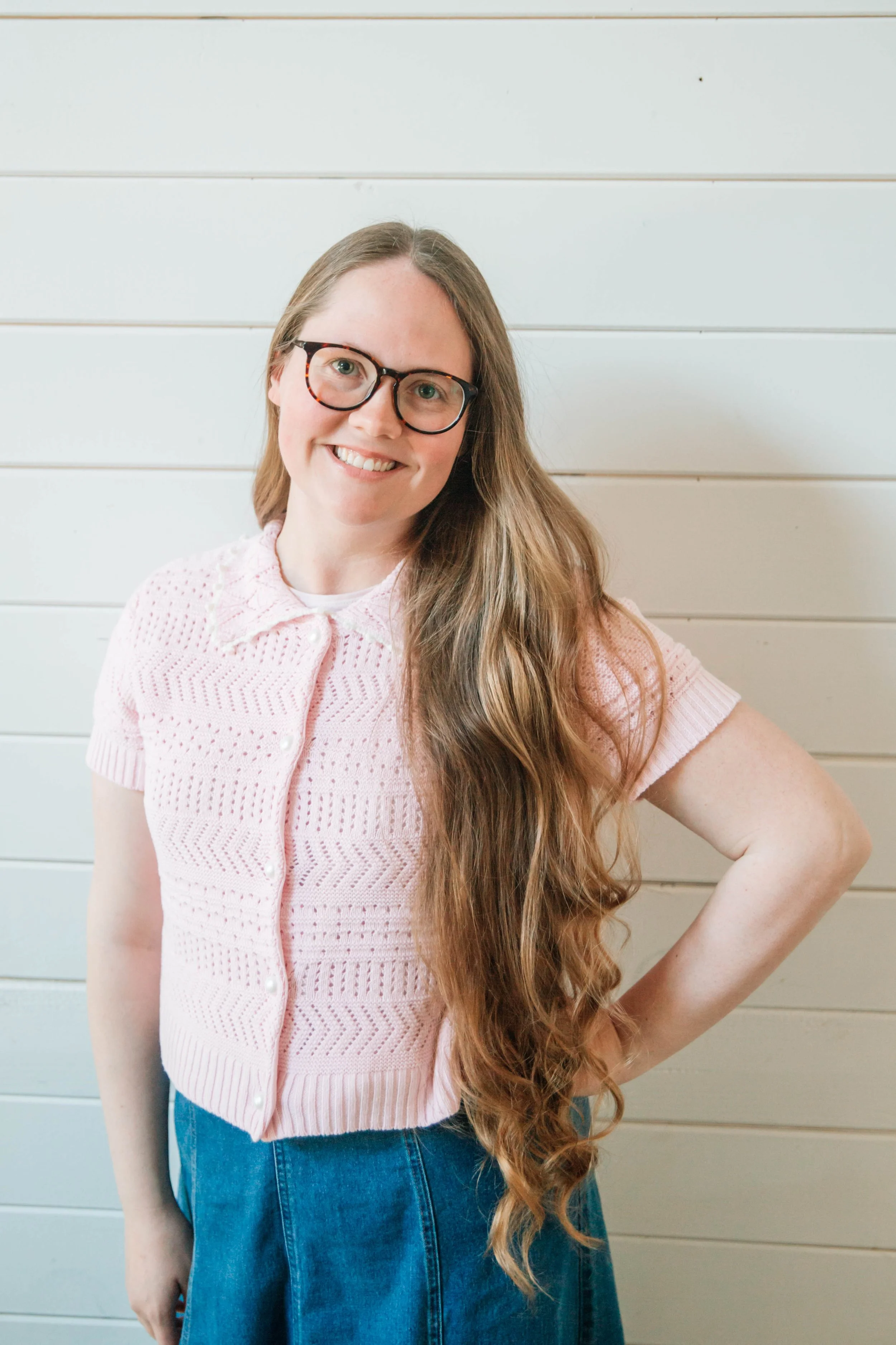 A young woman mortgage broker with long, wavy hair wearing glasses and a pink knitted short-sleeve top, smiling and standing against a light-colored wooden wall thinking about mortgages, preapproval, refinance, renewal.