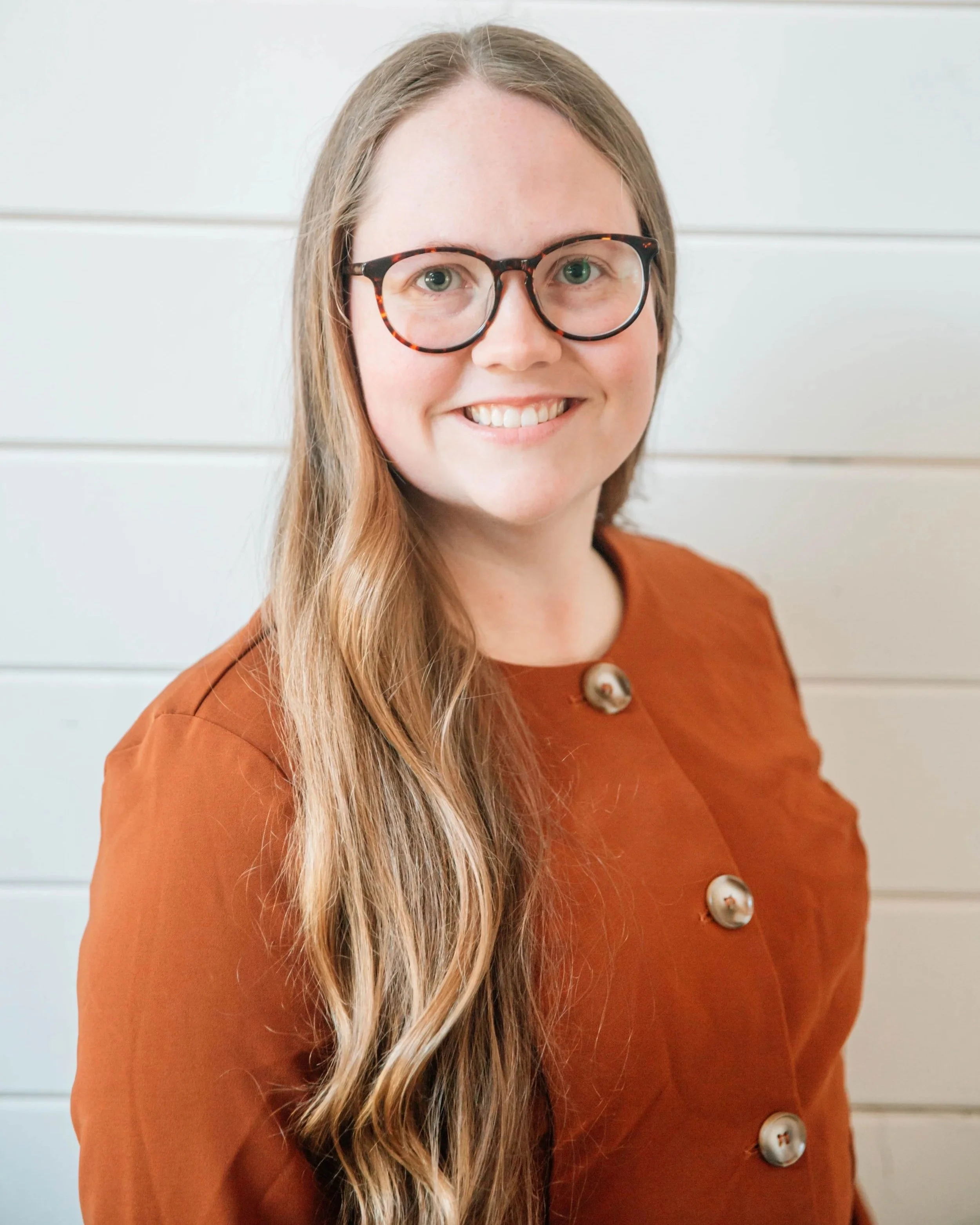 Close-up of a woman mortgage broker with long blonde hair, wearing tortoiseshell glasses and an orange-brown top with large buttons, standing against a white horizontal panel background thinking about mortgages.