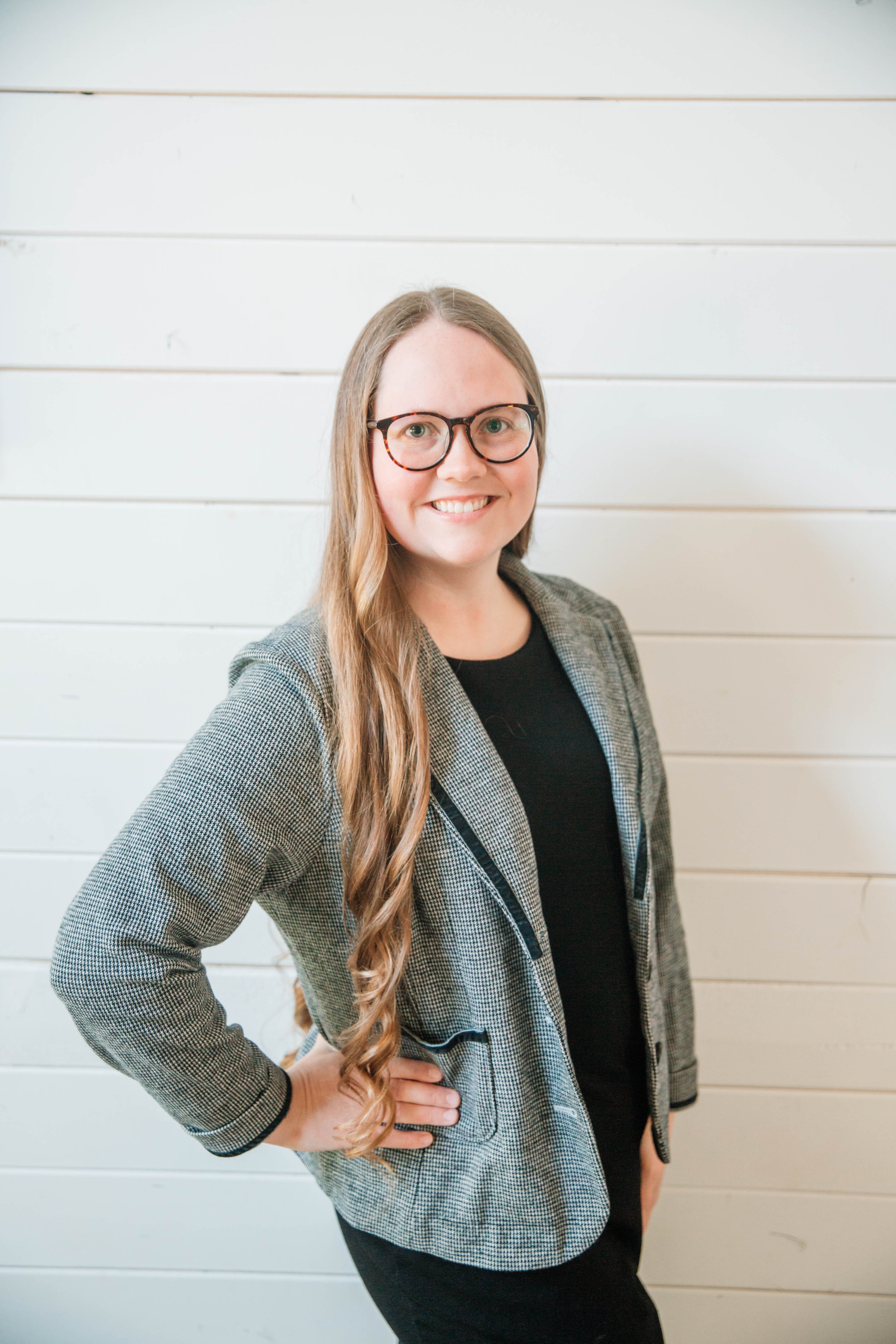 A woman mortgage broker with long blonde hair, glasses, and a checkered blazer smiling and posing with her hand on her hip thinking about mortgages in front of a white wooden wall.