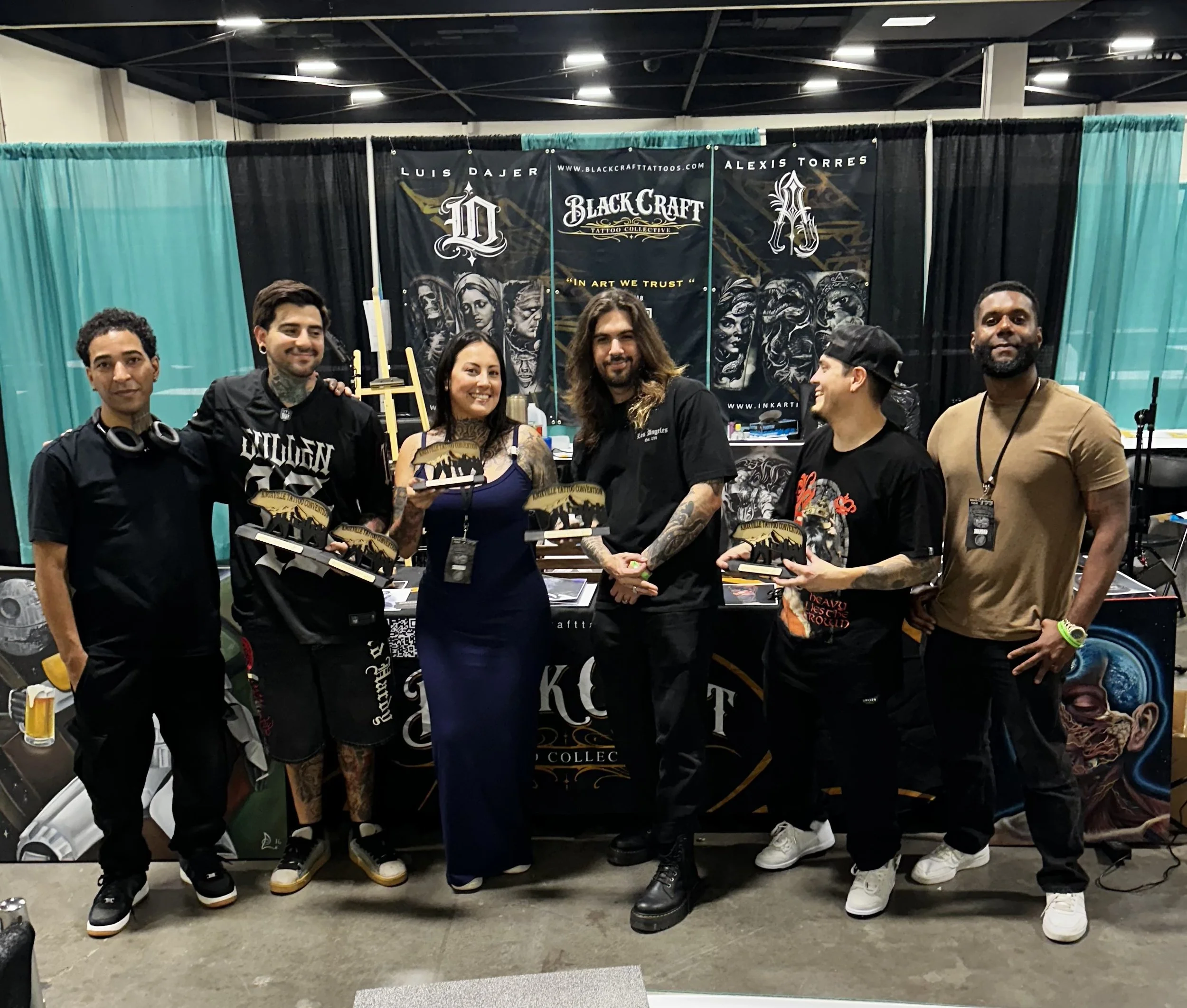 A group of five people standing in front of a black curtain display at a tattoo convention. The display features banners with text and graphics promoting Black Craft Tattoo Collective and its artists. The people are smiling and holding awards or trop