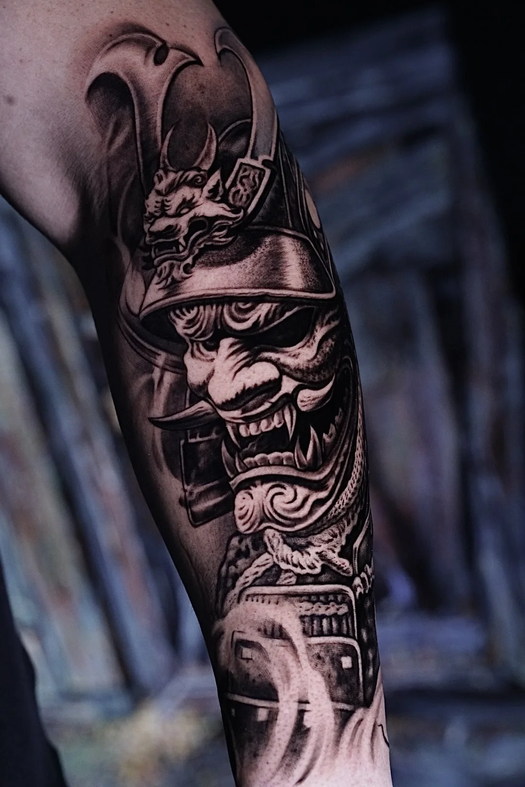 Black-and-gray demon mask and dice tattoo on the arm at Black Craft Tattoo Collective.