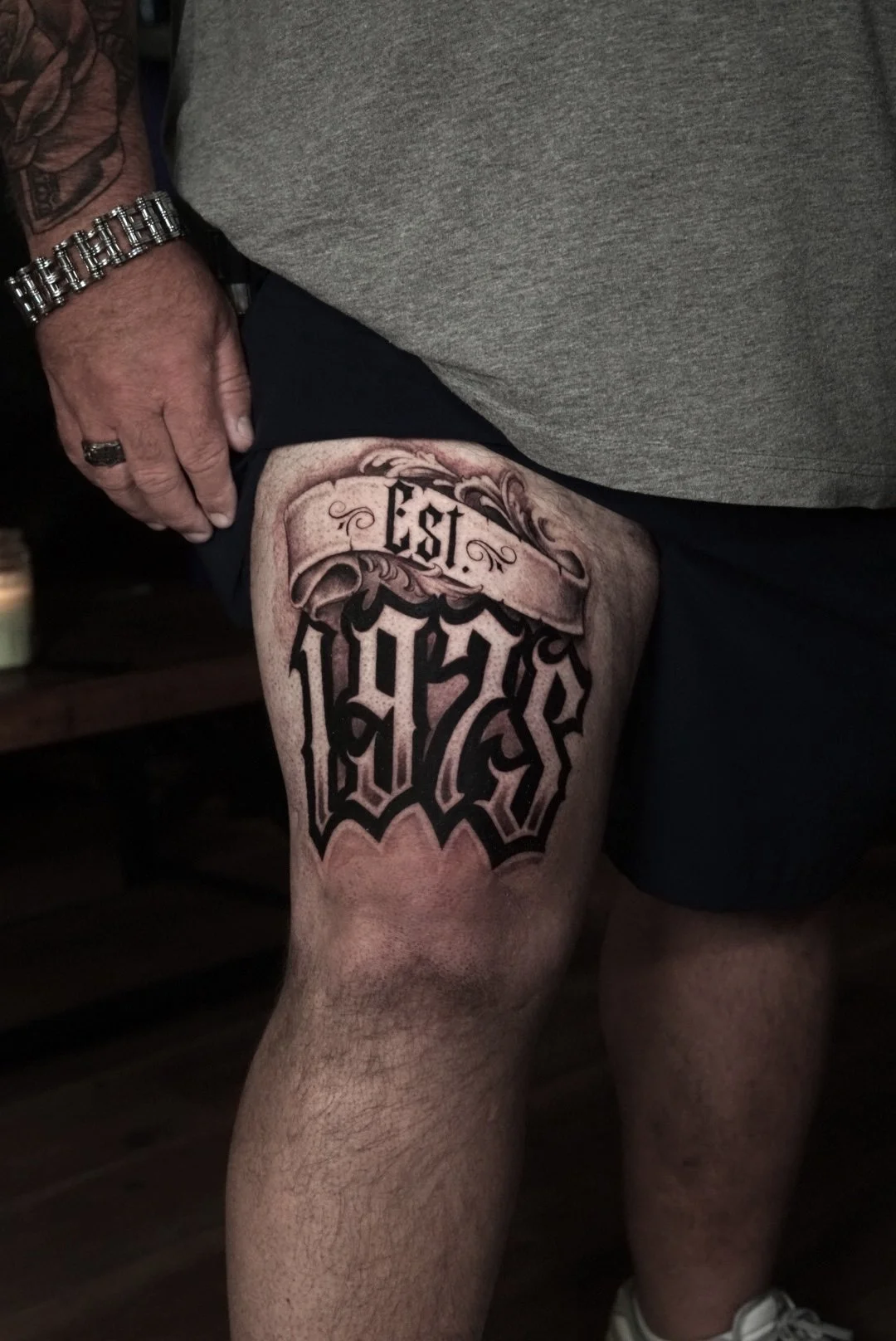 Blackletter Established 1984 thigh tattoo with ornamental scrollwork at Black Craft Tattoo Collective.