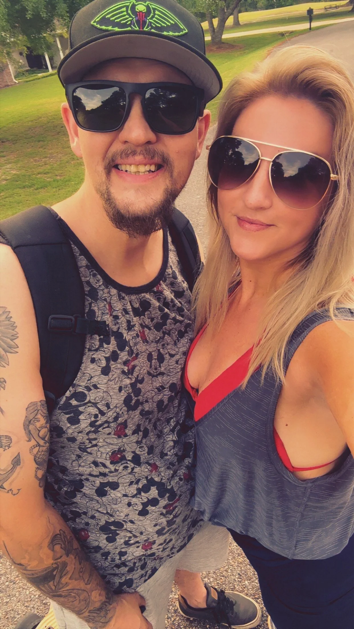A smiling man wearing a black cap with a glowing green insect design, black sunglasses, a sleeveless shirt with a pattern, and tattoos on his arm. A woman wearing large sunglasses and a gray shirt with a red strap is standing close to him. They are outdoors on a sidewalk with a grassy area and trees in the background.