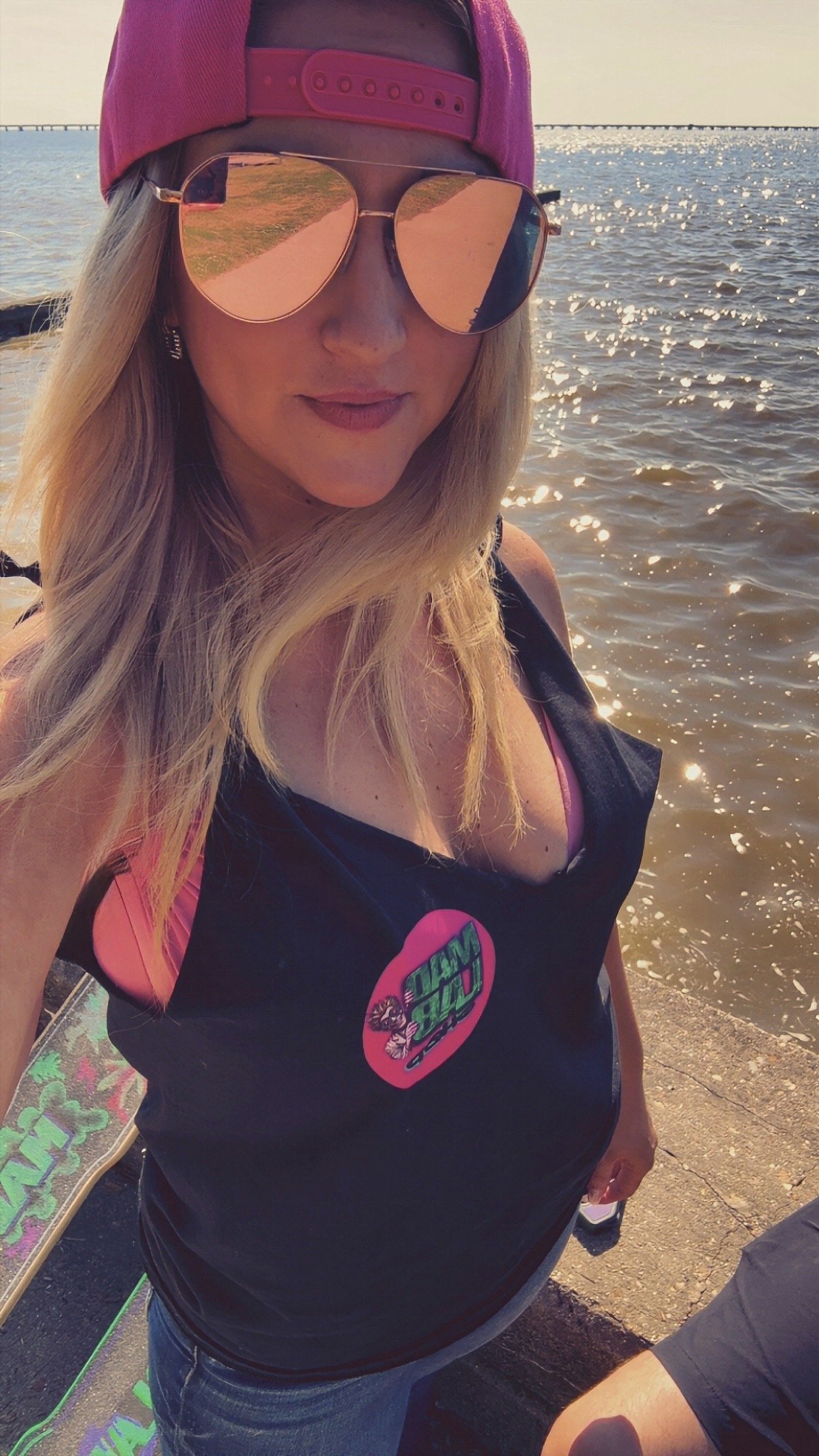 A woman with long blonde hair wearing mirrored sunglasses and a pink baseball cap takes a selfie near the water. She is dressed in casual clothes with a pink sports bra visible underneath a black shirt that has a colorful logo. The reflection in her sunglasses shows the water and sky. The background features water with sunlight reflecting off it and a bridge or pier in the distance.