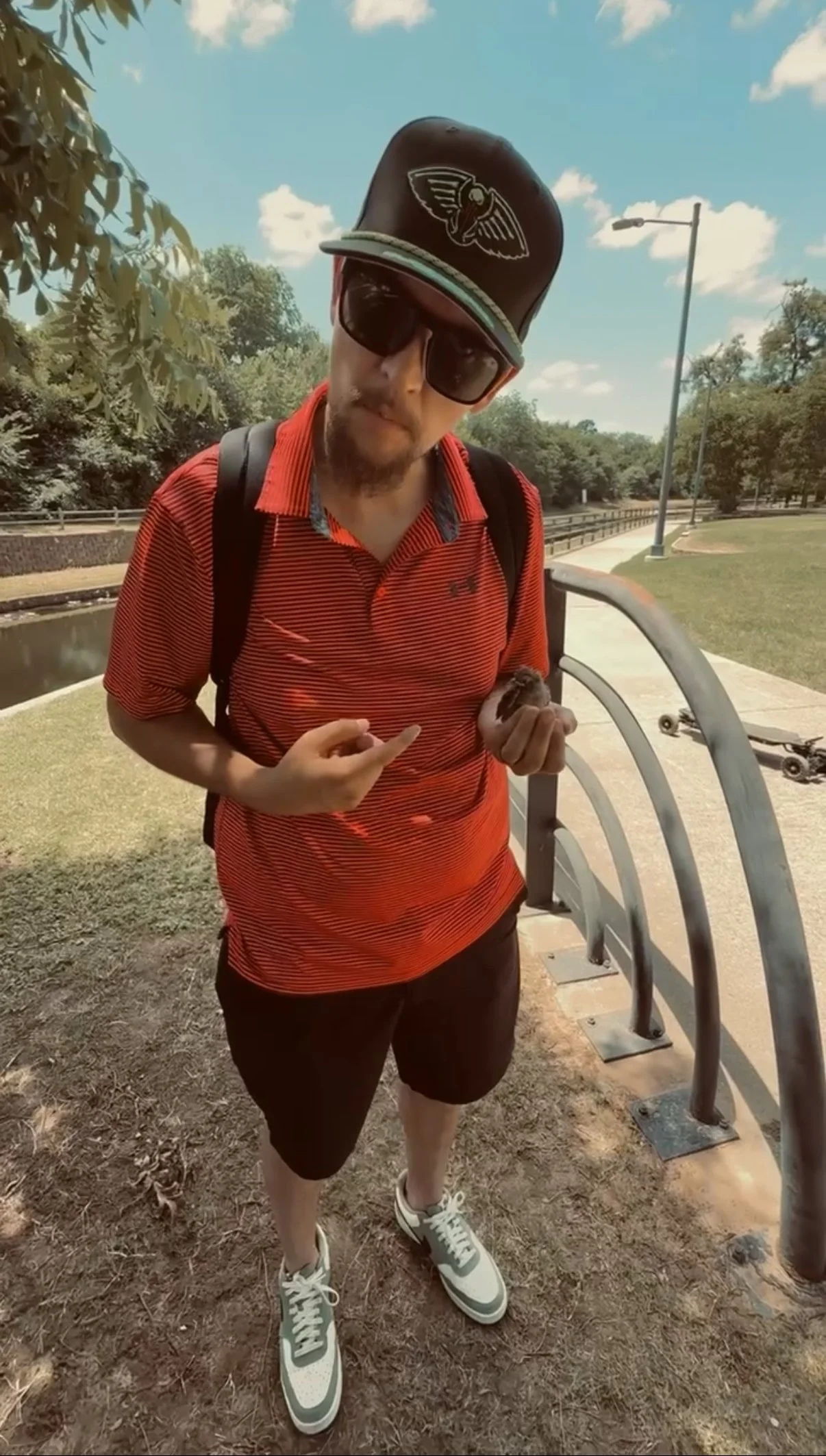 A man wearing sunglasses, a black cap with a logo, a red striped polo shirt, black shorts, and sneakers, standing outdoors on a dirt path near a park railing with trees and a skateboard on the ground nearby.