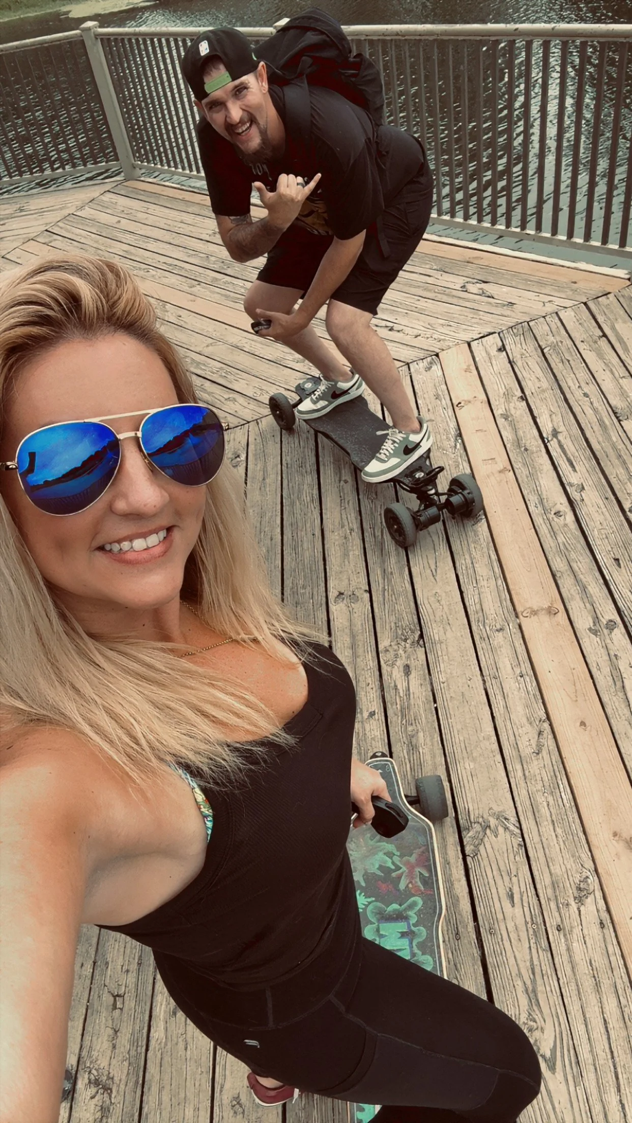 A woman with blonde hair wearing sunglasses and a black tank top, taking a selfie on a wooden dock near a body of water, with a skateboard beside her. A man making a silly face and gesture is on a skateboard in the background.