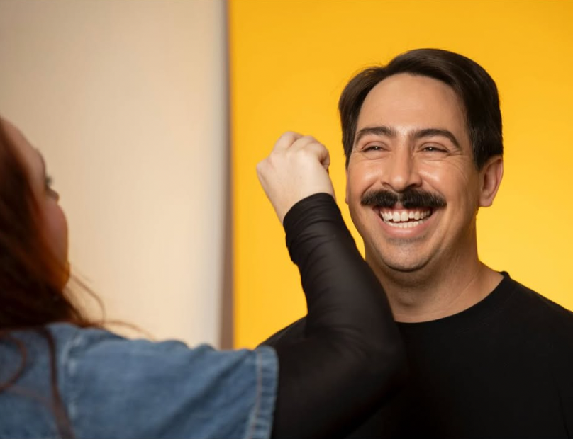 Person applying makeup or face paint to a smiling man's face, with a bright yellow background.