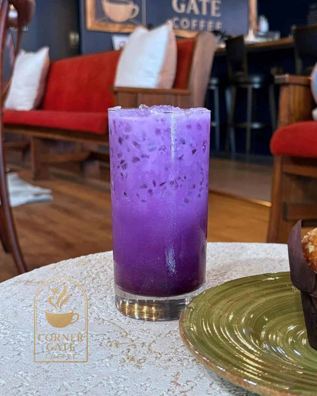 A tall glass of purple iced beverage with ice cubes on a table in a coffee shop. In the background, there are red upholstered benches, white pillows, a chalkboard sign, and wooden chairs and tables.