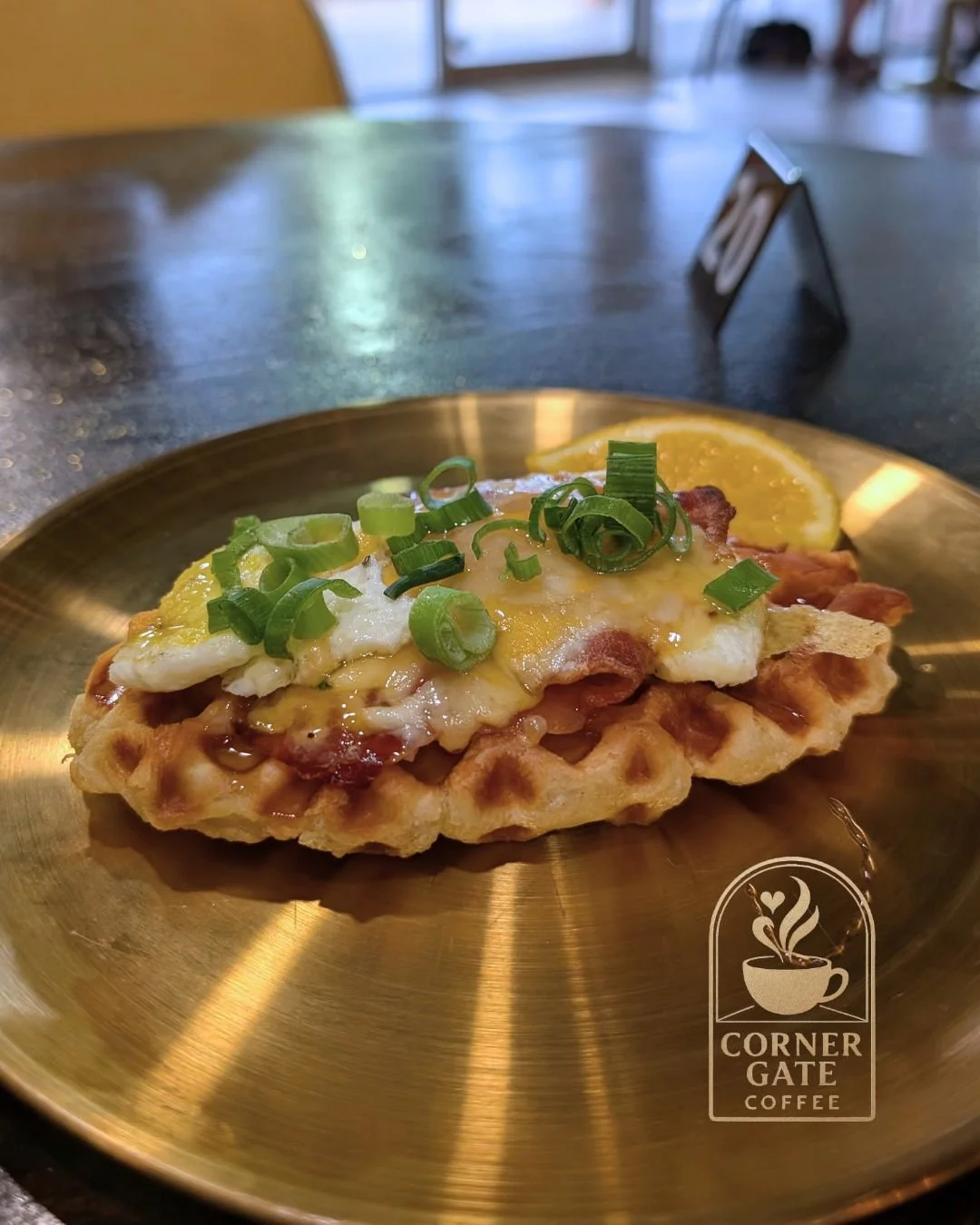 Waffle topped with fried egg, green onions, bacon, and lemon wedge on a gold plate at Corner Gate Coffee.