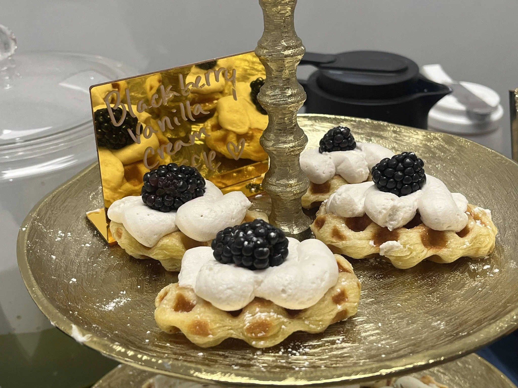 A tiered dessert display with small waffle base topped with whipped cream, blackberries, and a decorative sign that reads 'Blackberry vanilla' in cursive font, set on a golden tray.