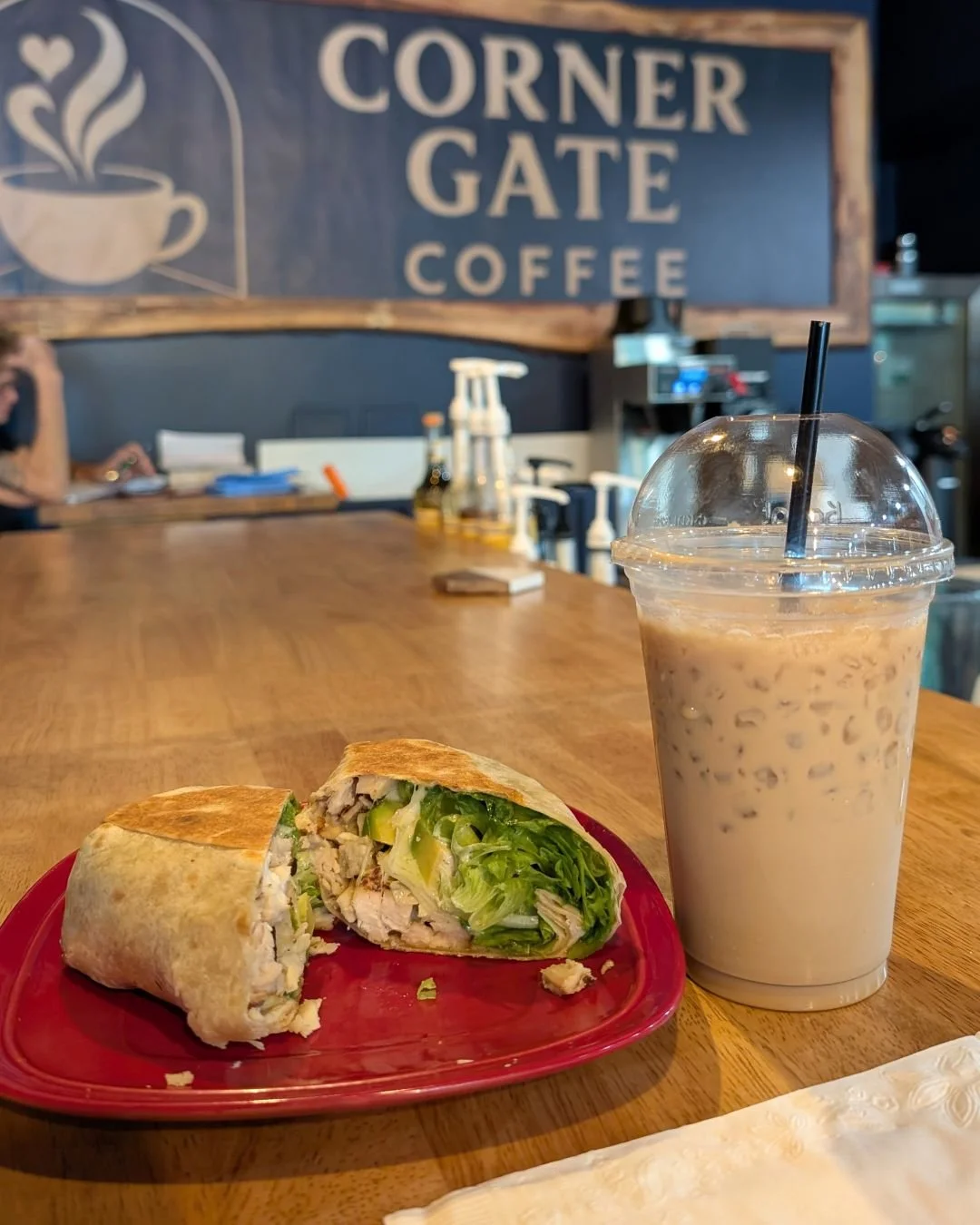A burrito cut in half on a red plate, with lettuce, chicken, and other ingredients inside, next to a large iced coffee in a clear plastic cup with a black straw, on a wooden table inside a coffee shop.