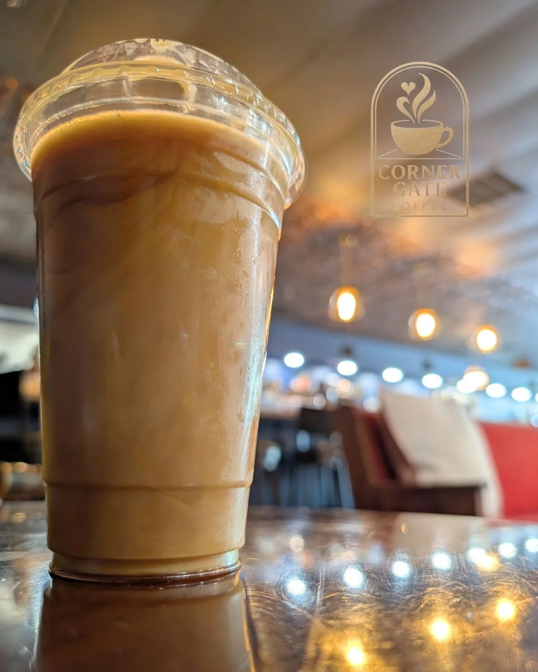 A close-up of a tall plastic cup filled with iced coffee on a shiny wooden table, with blurred indoor background and a logo for Corner Gate Coffee visible on the window.