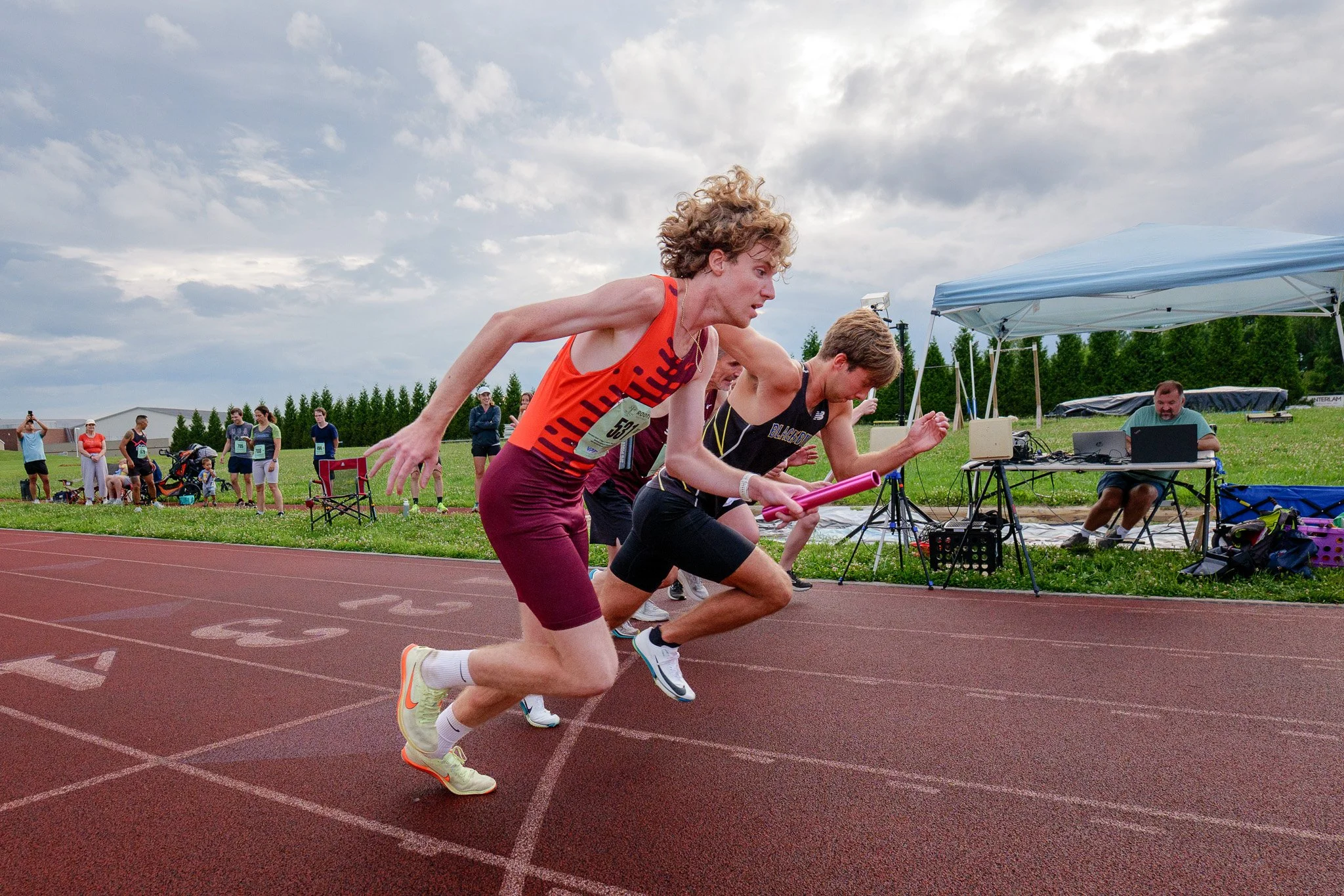 Two male athletes are sprinting on a red running track during a race, with several spectators watching in the background and a person at a table with electronic equipment under a canopy.