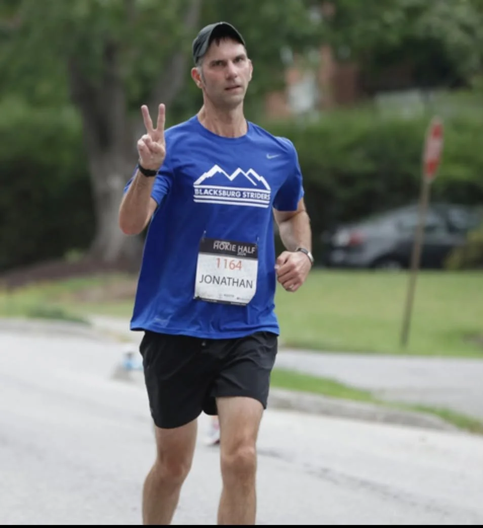A man running in a race, holding up a peace sign with his right hand, wearing a blue shirt and black shorts, with a race bib that reads 'Jonathan' and a number, in a park or outdoor environment.