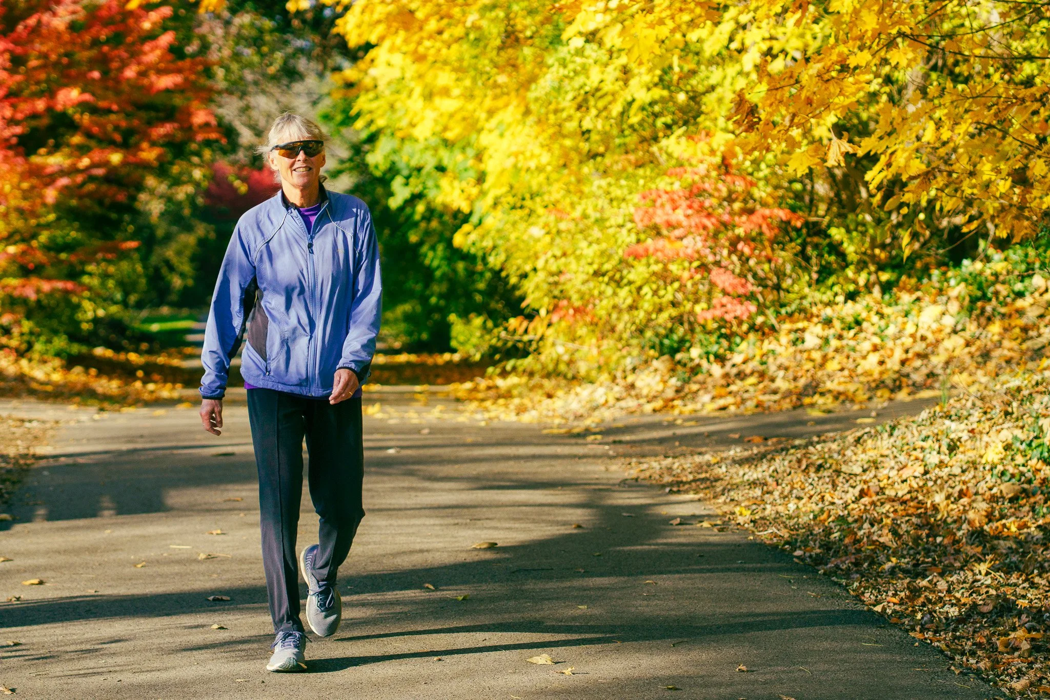 A woman walking on a paved path surrounded by vibrant fall foliage with yellow, orange, and red leaves, wearing sunglasses, a purple and blue athletic jacket, and gray pants.