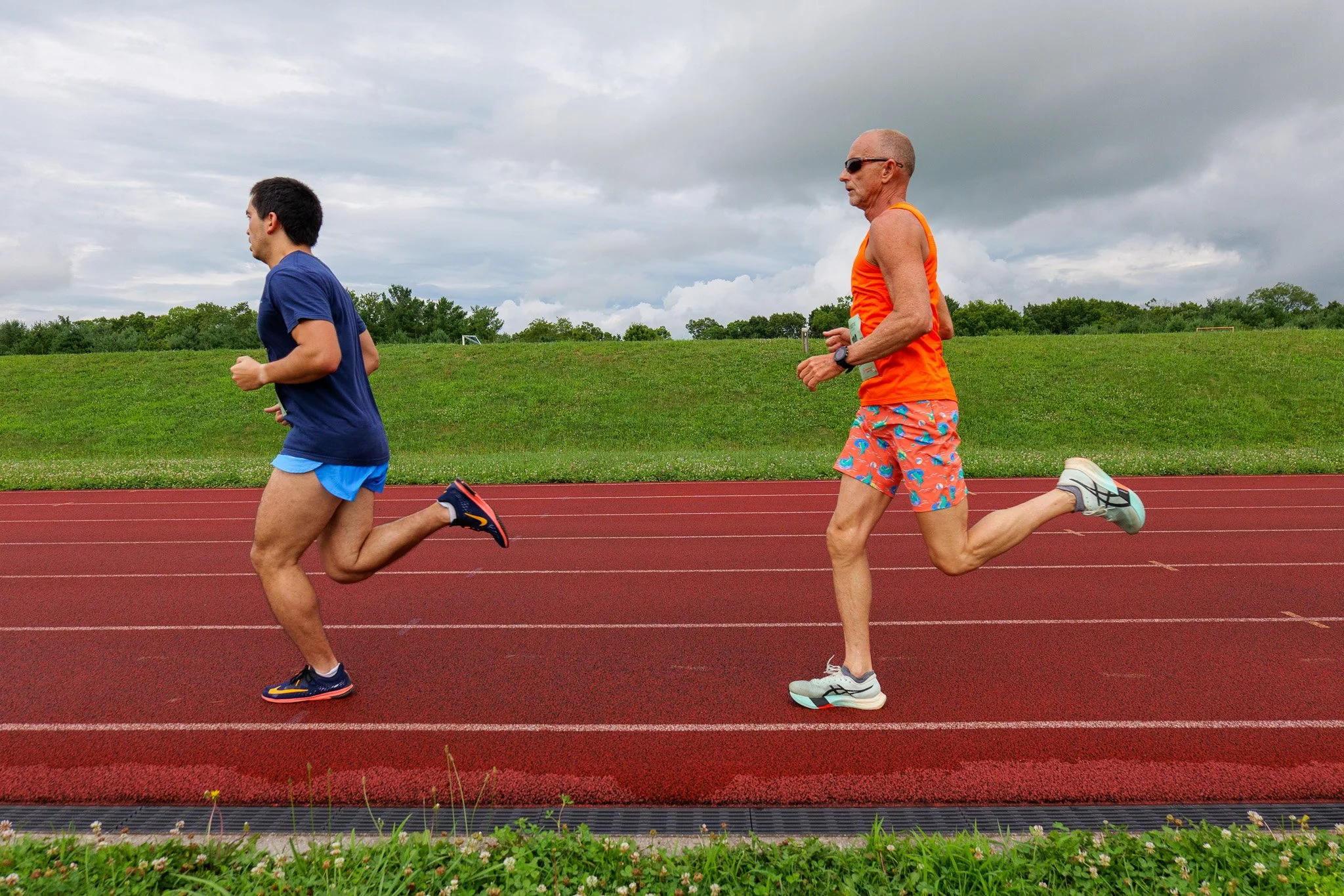 Two male runners, a younger man and an older man, running on a track outdoors on a cloudy day. The younger man wears a blue shirt and shorts, while the older man wears an orange tank top and colorful shorts.