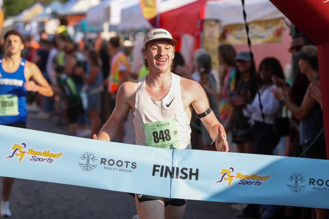 A male runner crossing the finish line at a race event, smiling with arms raised, wearing a white Nike tank top, a cap, and a race bib number 848, with onlookers and other runners in the background.