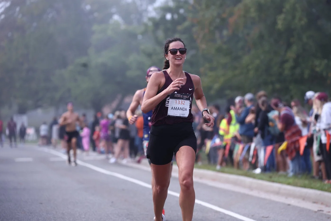 A woman running in a marathon race, wearing sunglasses, a black sports tank top, black shorts, and a race bib with the number 1332 and name Amanda. There are people watching and cheering on the side of the road, with trees in the background.