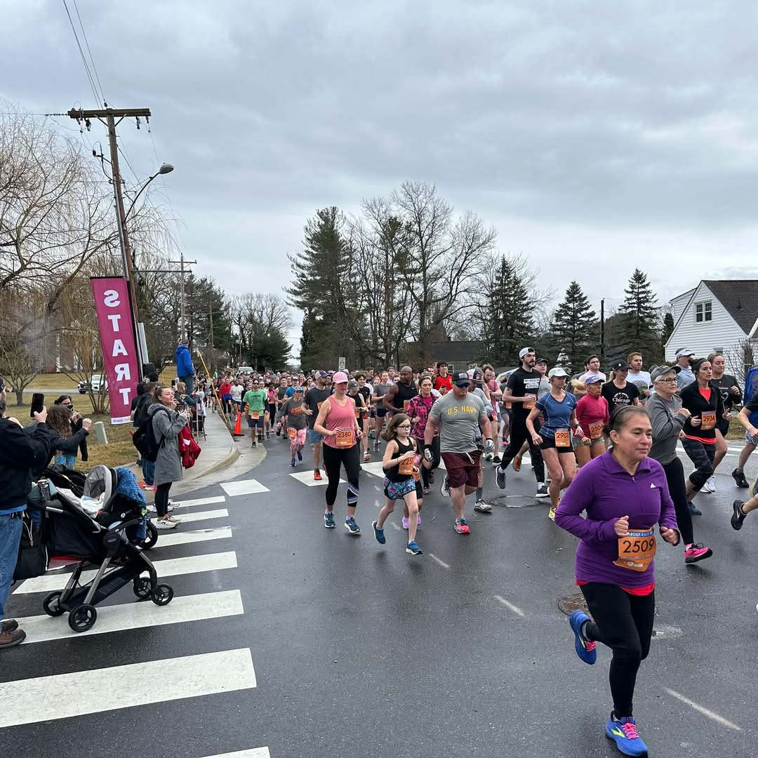 Group of people participating in a city marathon on a cloudy day, with some spectators taking photos. Runners wearing athletic clothes and numbered bibs cross a street with a 'START' banner on the sidewalk.