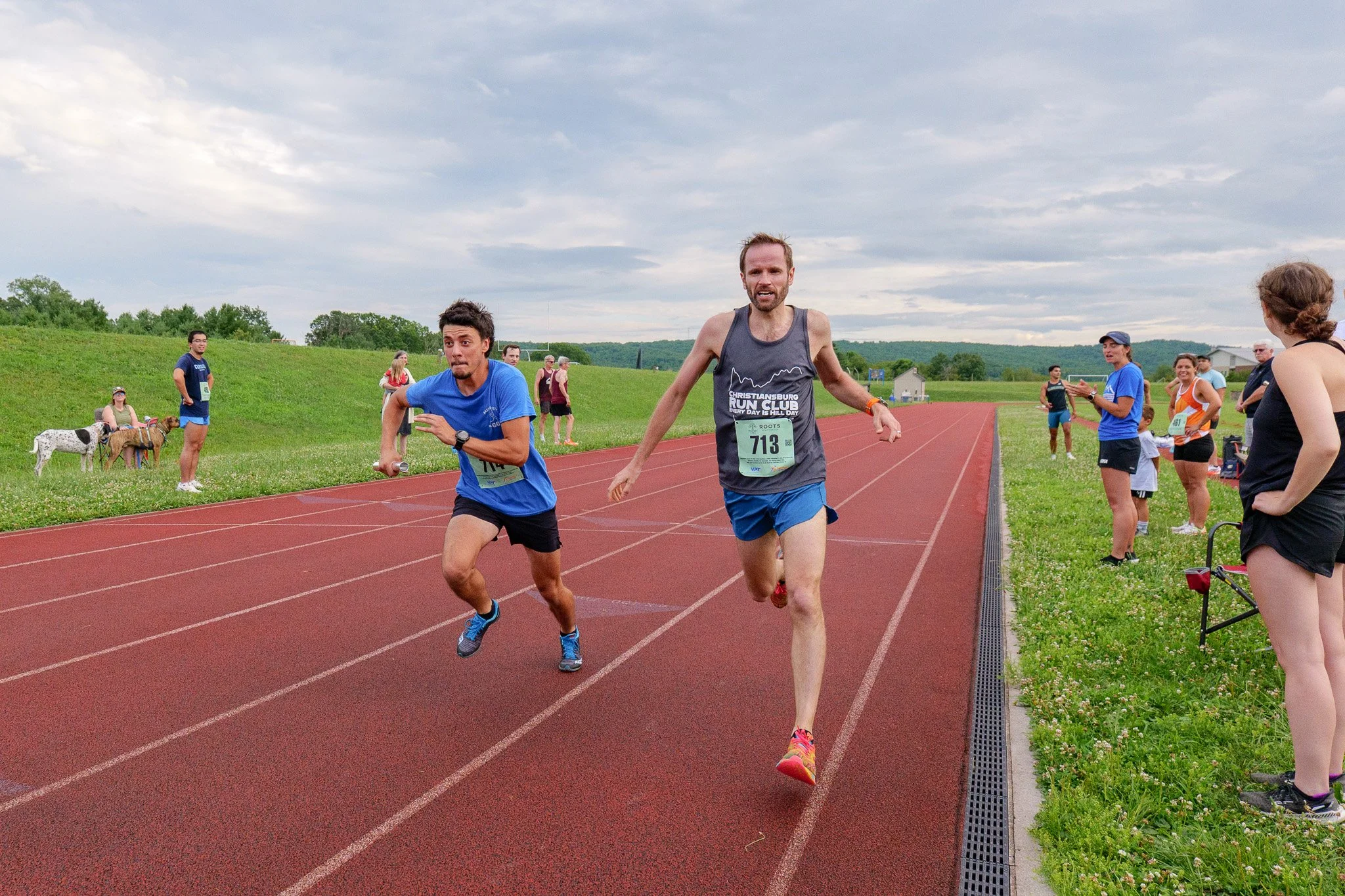 Two male runners competing in a race on an outdoor track, with spectators and dogs observing on the grassy side, under a cloudy sky.