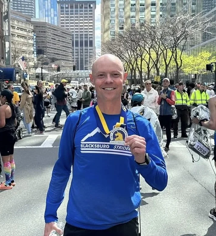 Man in blue marathon shirt holding a medal at a city race with spectators and tall buildings in the background.
