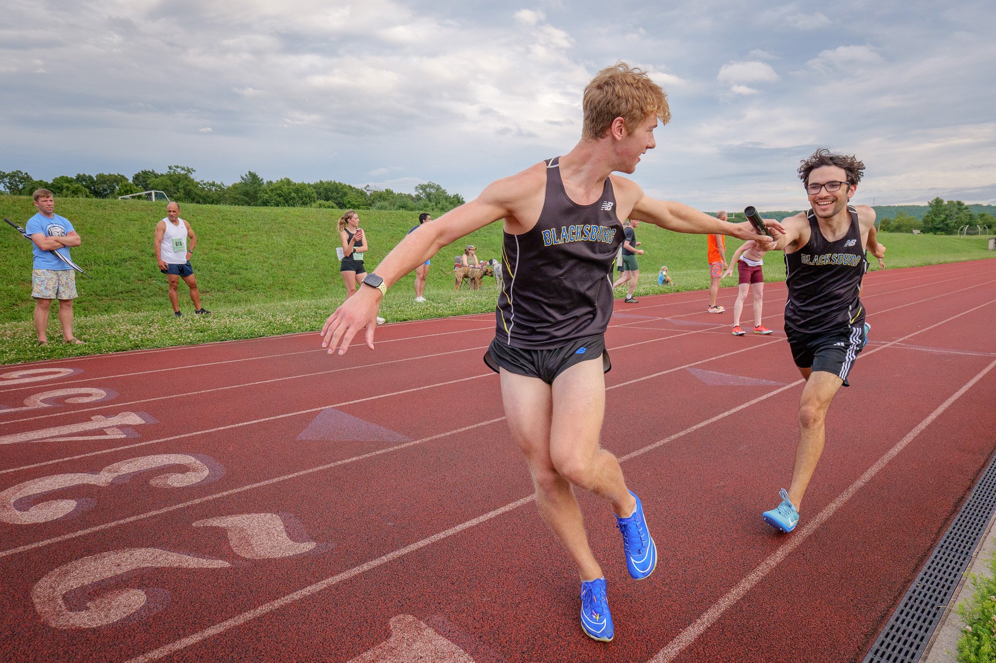 Two male runners in black athletic gear with 'BLACKSBURG' written on their shirts are in the middle of a race on a red track, smiling and reaching out to high-five each other as they finish.