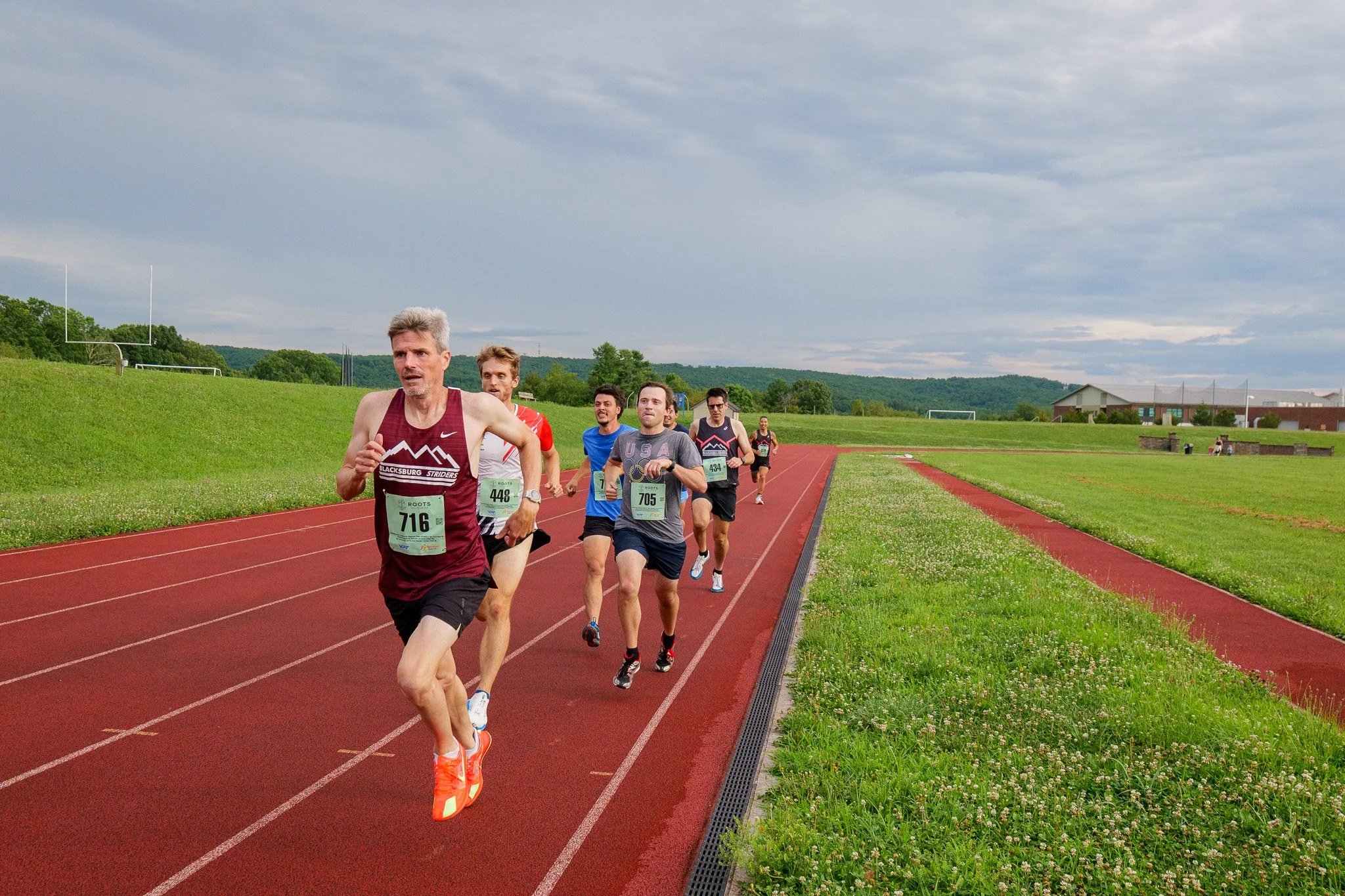 A group of six men running on an outdoor track field with green grass on either side. Overcast sky with hills and buildings in the background.