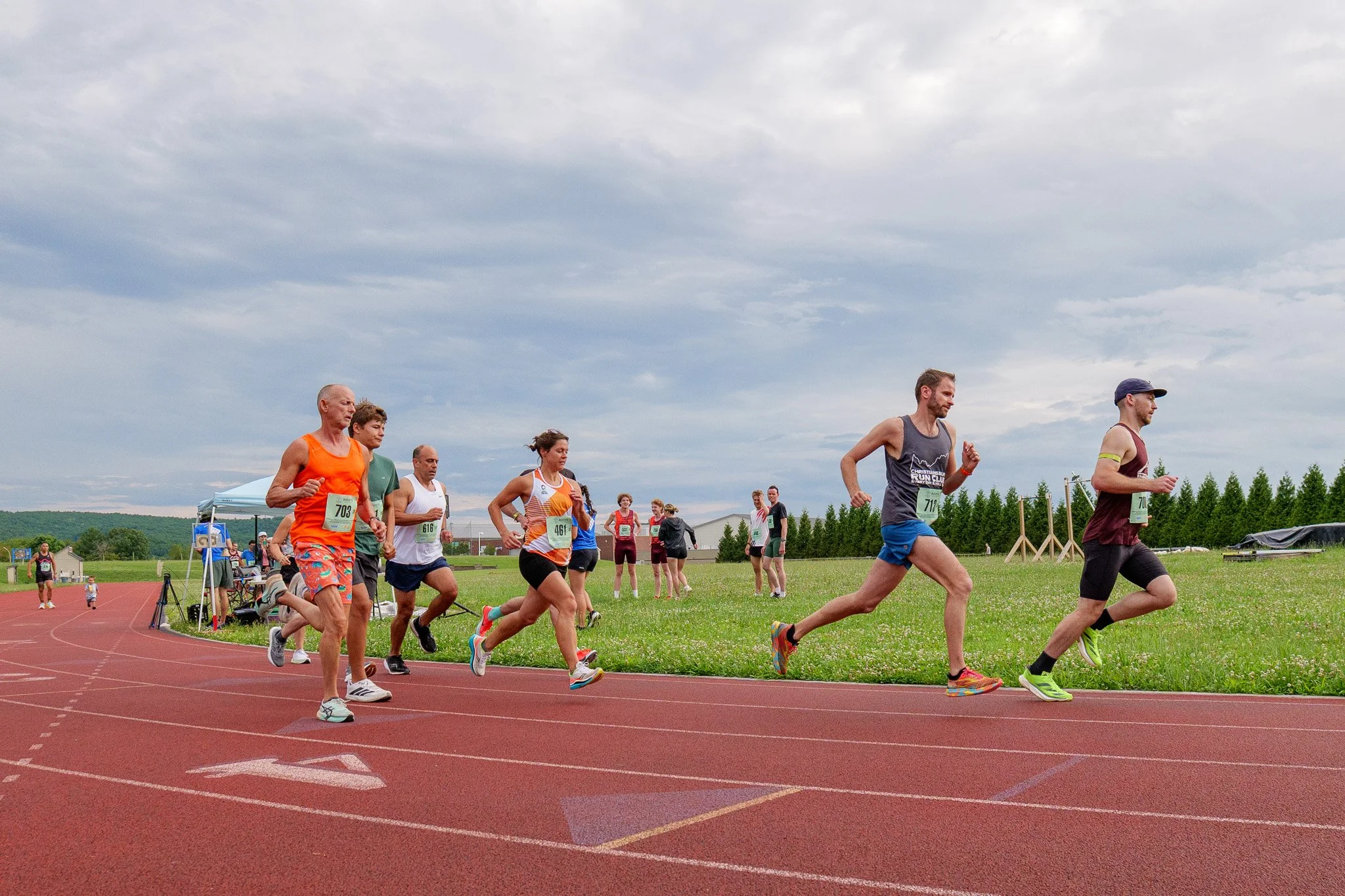Group of runners participating in a race on a track, with some people running and others walking, under a cloudy sky, surrounded by green fields and trees.