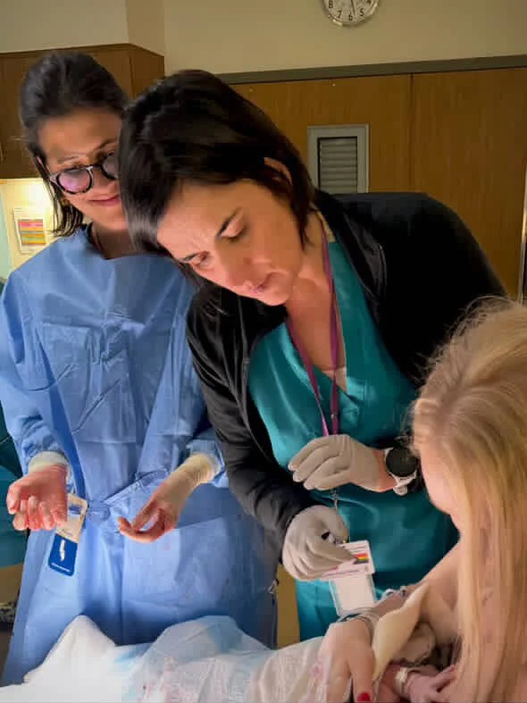 Two healthcare professionals, one wearing blue scrubs and another wearing teal scrubs, attend to a patient in a hospital room. The professional in teal appears to be performing a medical procedure on a young girl with blonde hair. The room has wooden paneling and a clock on the wall.