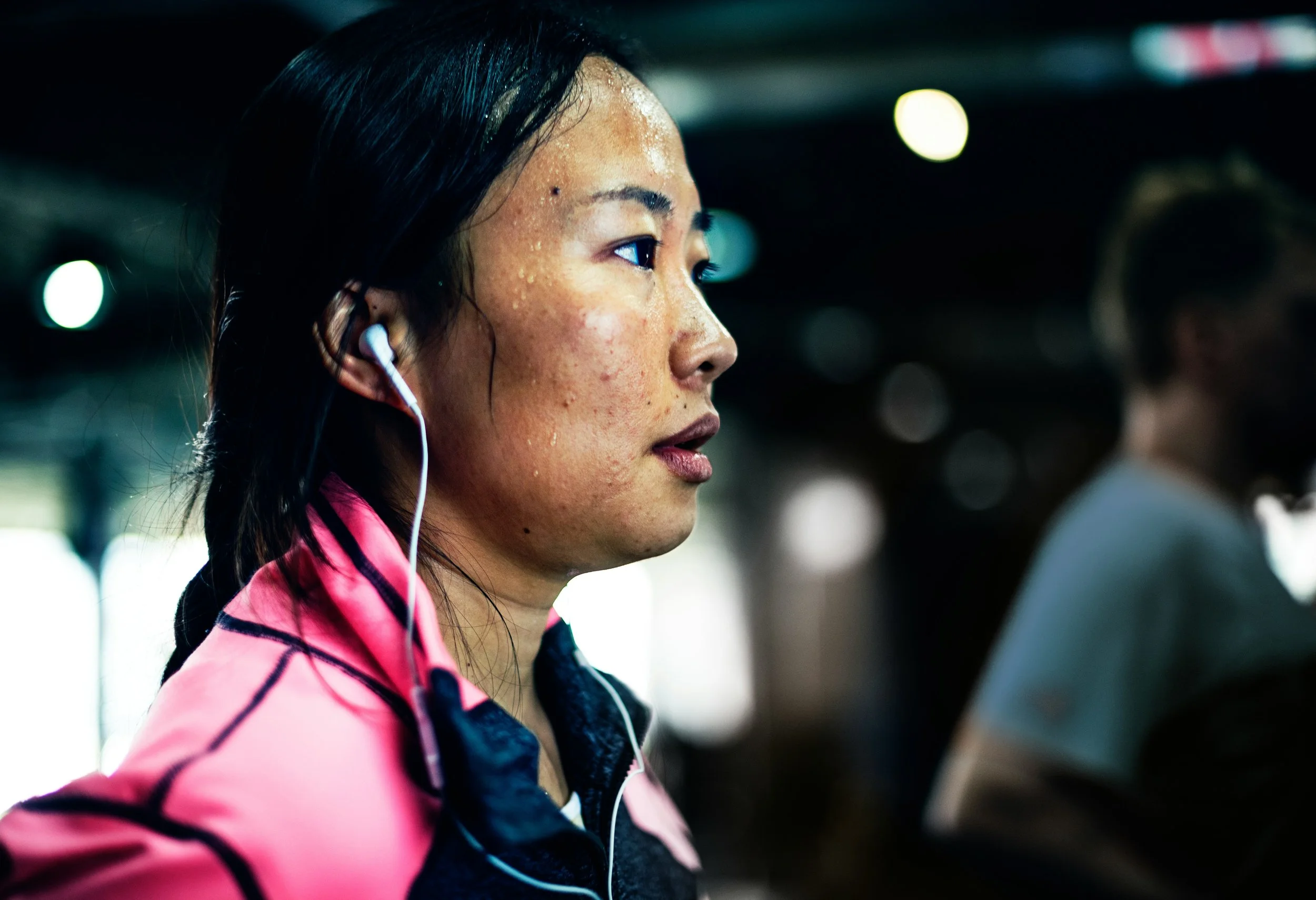 A woman with sweat on her face, wearing pink and black athletic clothing, listening to music with white earphones, and focusing on something in a gym or fitness area.