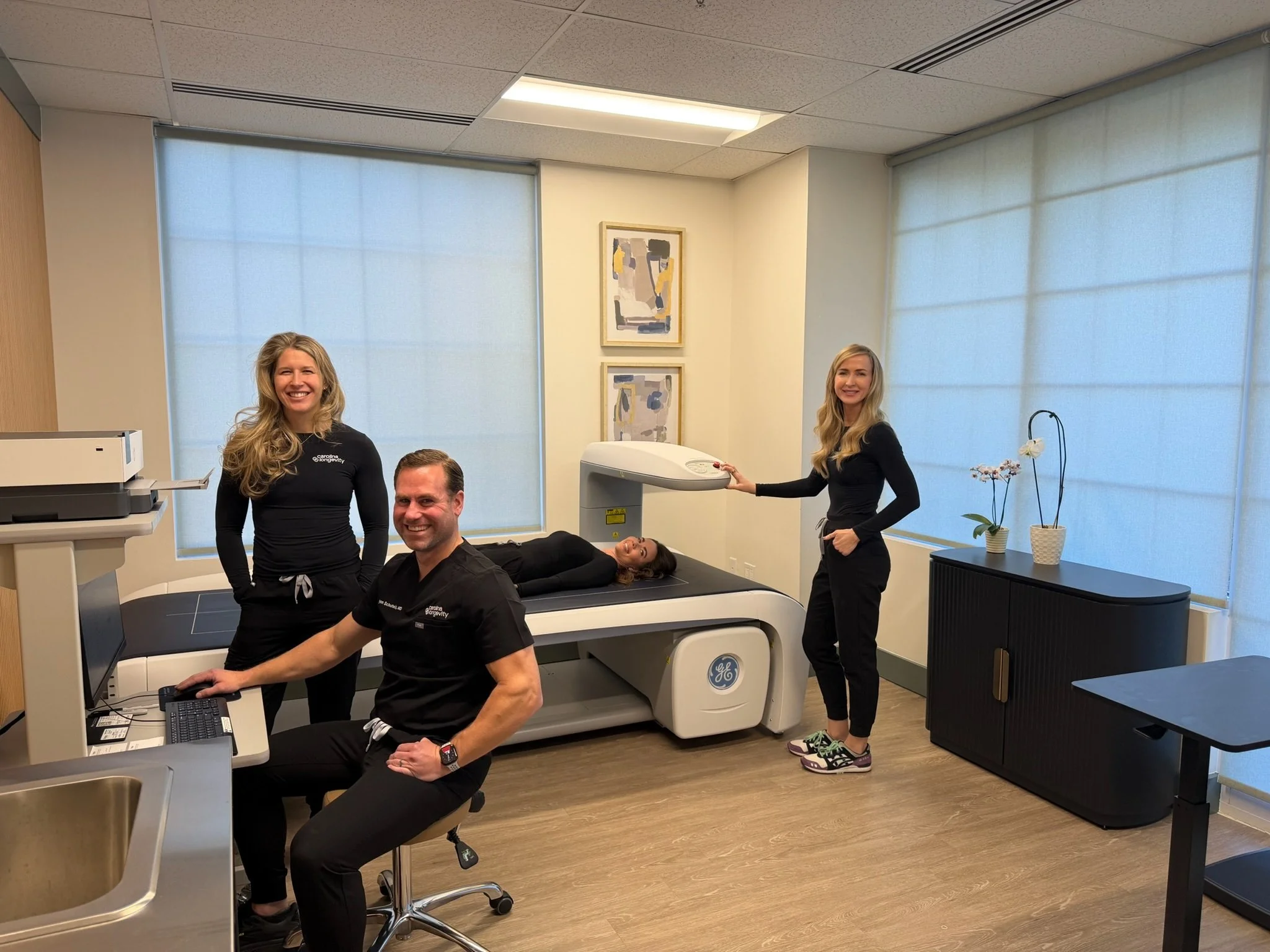 Medical imaging room with four people. One person is lying on an MRI or CT scan machine. Two women are standing near the scan, and a man is seated at a computer. The room has natural light with large windows covered with light blinds, and wall art and potted plants are visible.