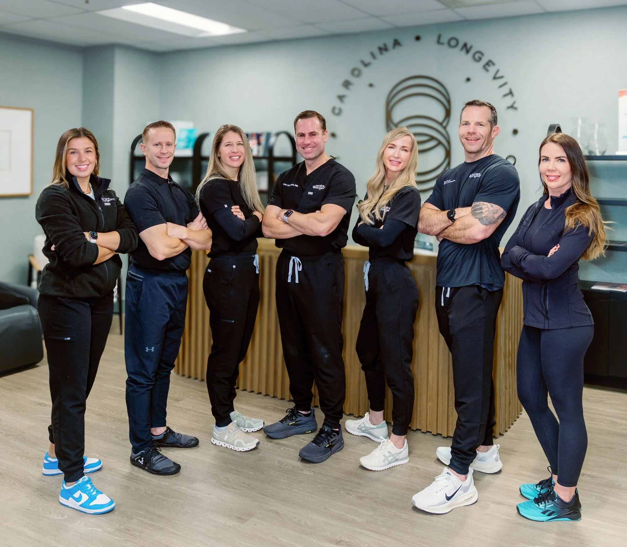 Group of seven fitness or healthcare professionals standing with arms crossed in an indoor office or clinic reception area.