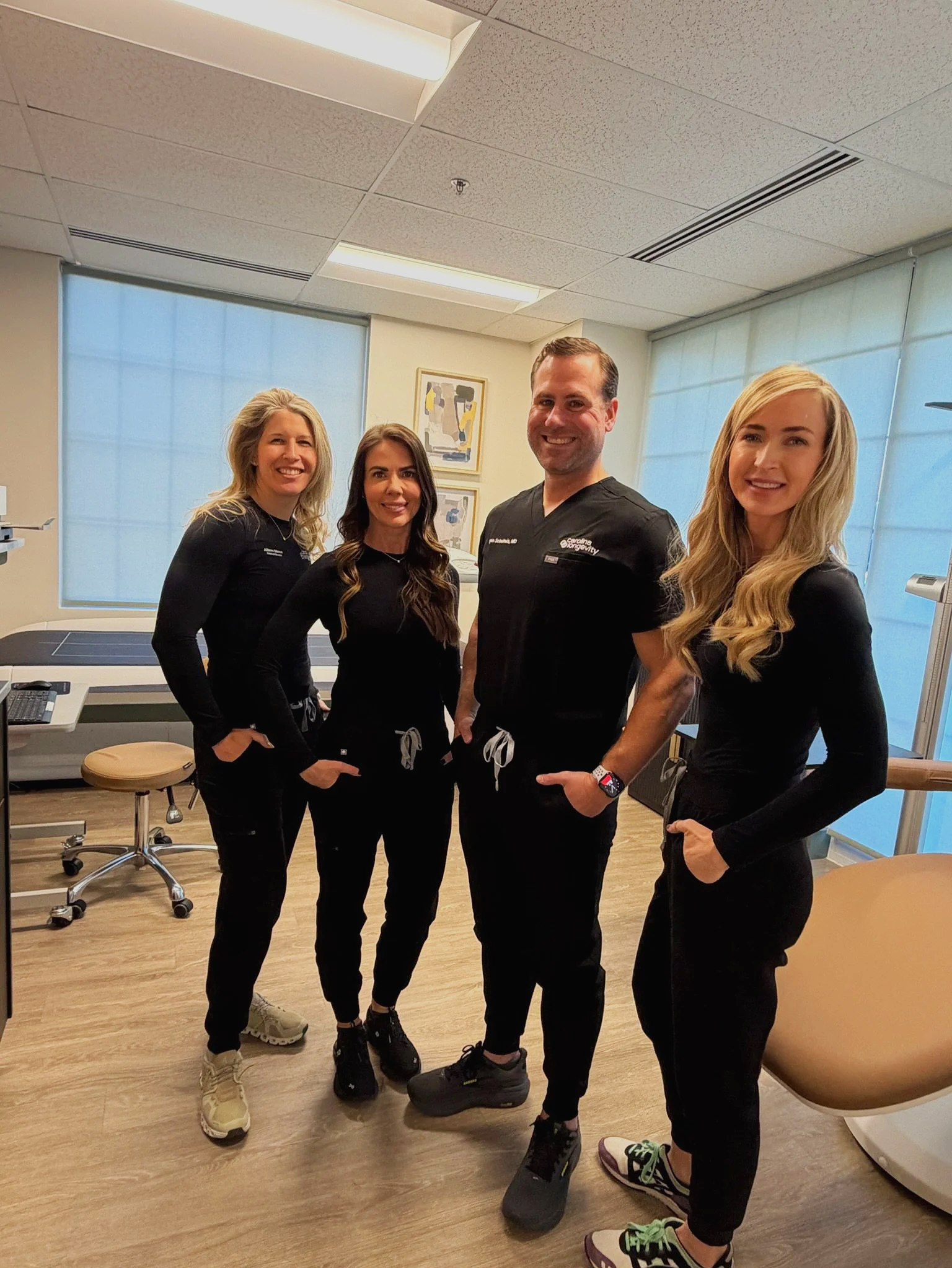 Four medical professionals, three women and one man, standing in a medical office, all wearing black scrubs and smiling at the camera.