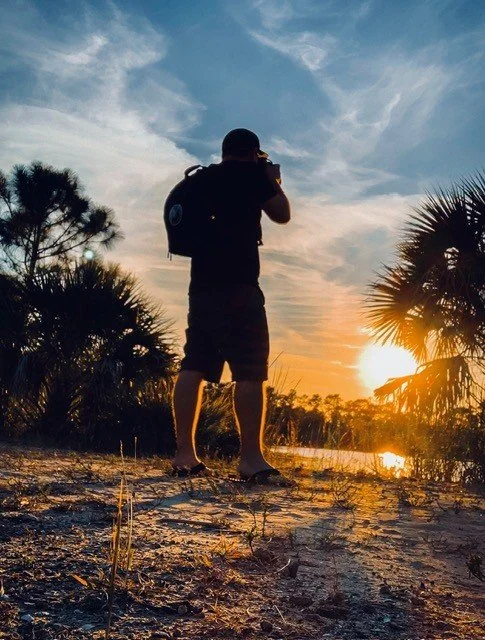 A man stands on a sandy path taking photos of a sunset with palm trees and water in the background.