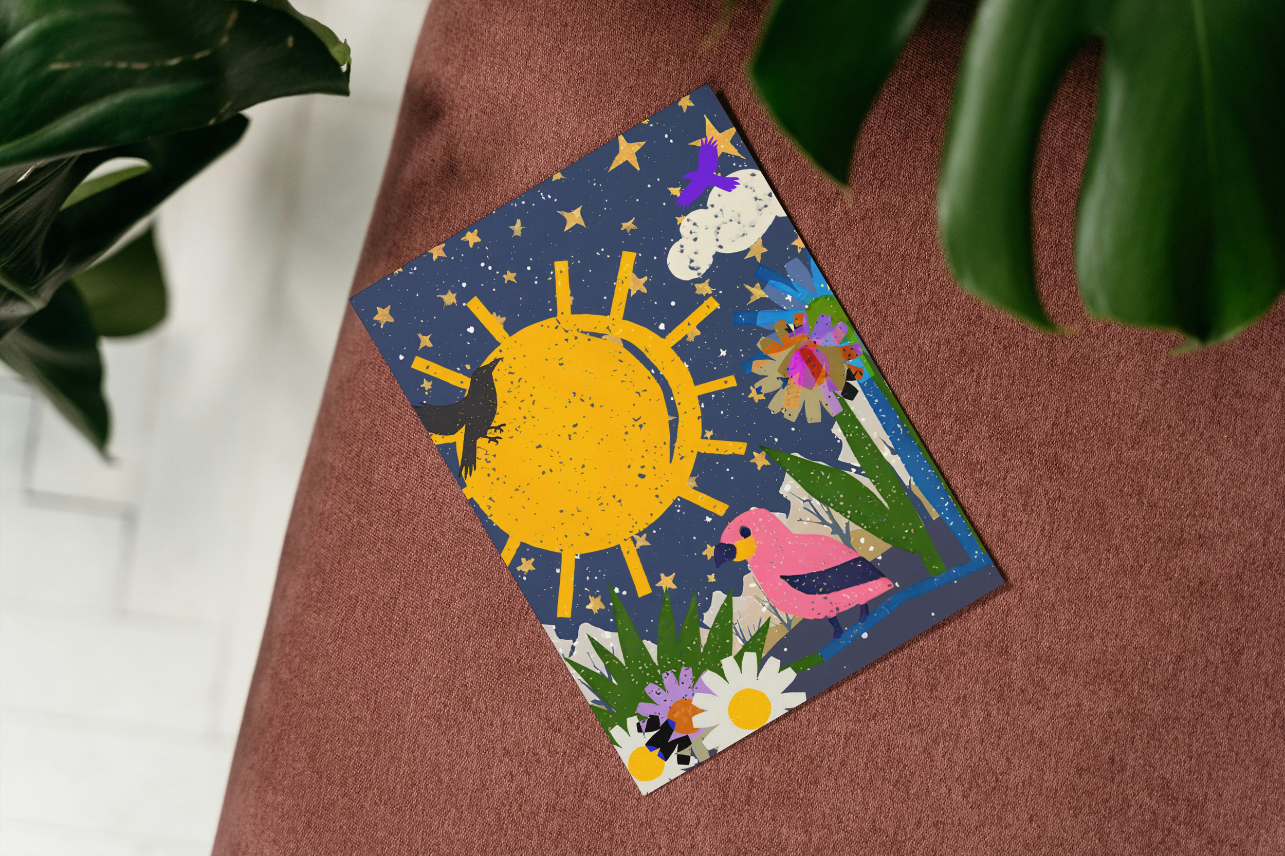 A colorful illustrated card with a sun, stars, a moon, a pink flamingo, a purple owl, flowers, and plant imagery, placed on a reddish-brown fabric surface, partially obscured by green leaves.