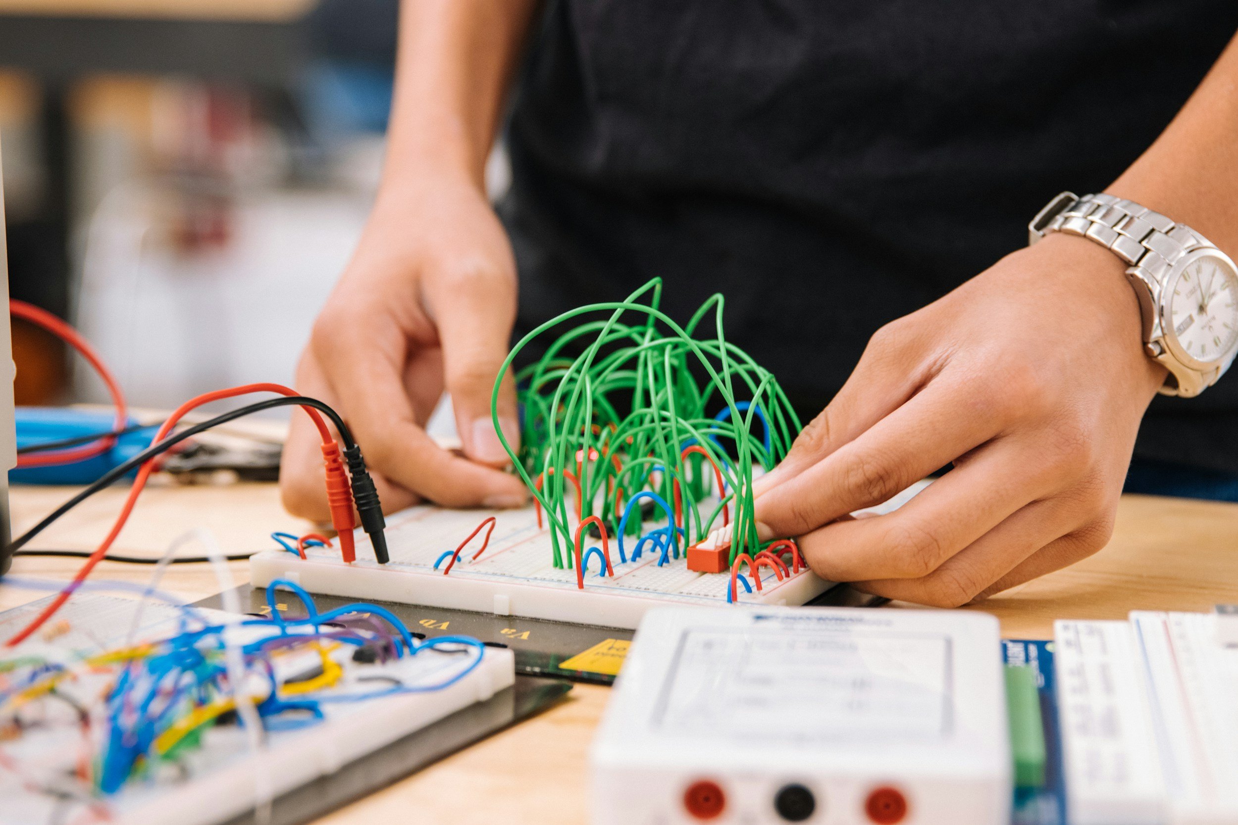 Person working on electronic breadboard circuit with colorful jumper wires and wires connected to various components, wearing a silver watch on their left wrist.