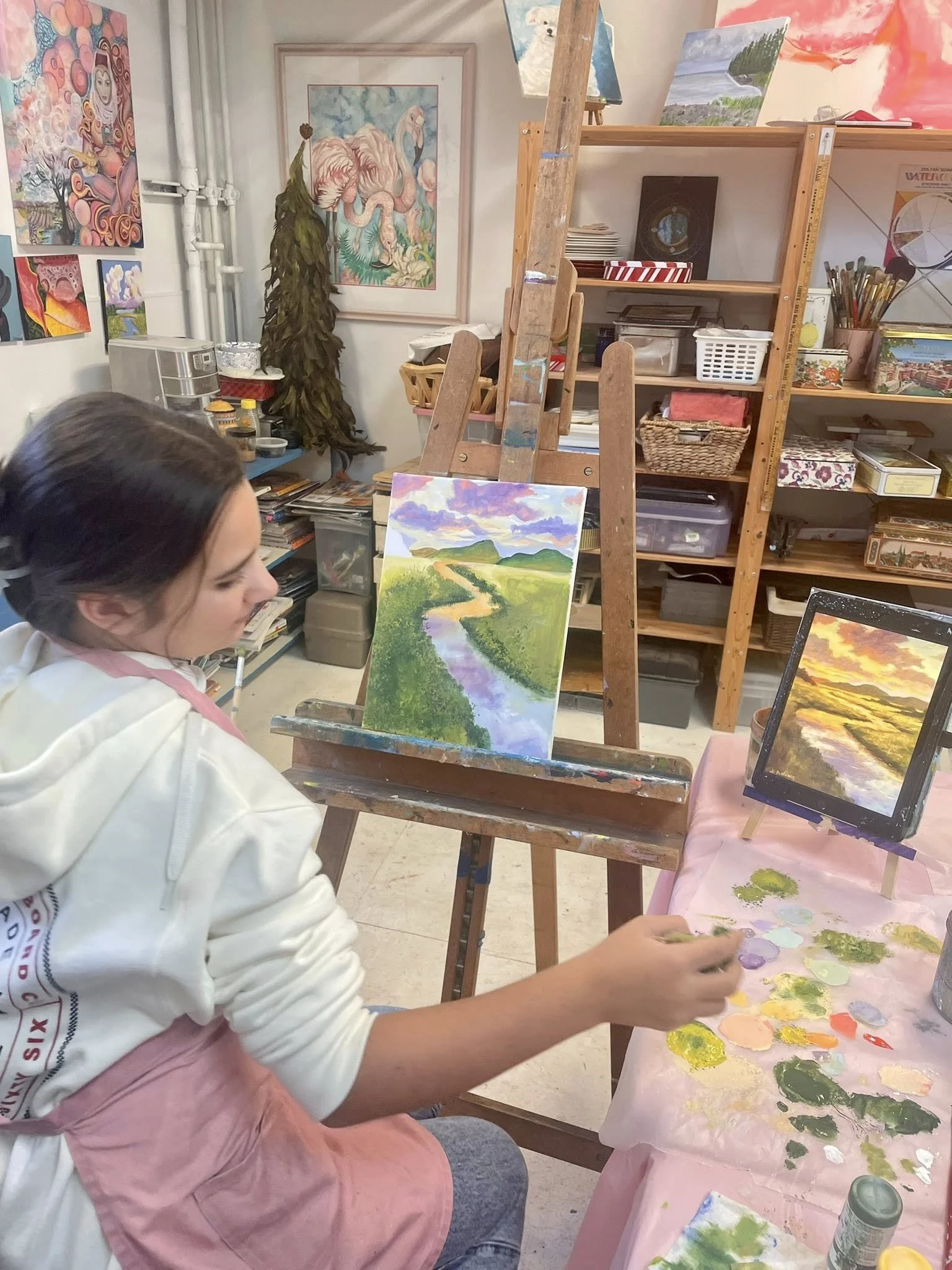Teen Student at Art from the Heart Ojai painting landscape scenery with a river, grassy hills, and a colorful sky. Art supplies and paintings are visible in an art studio.