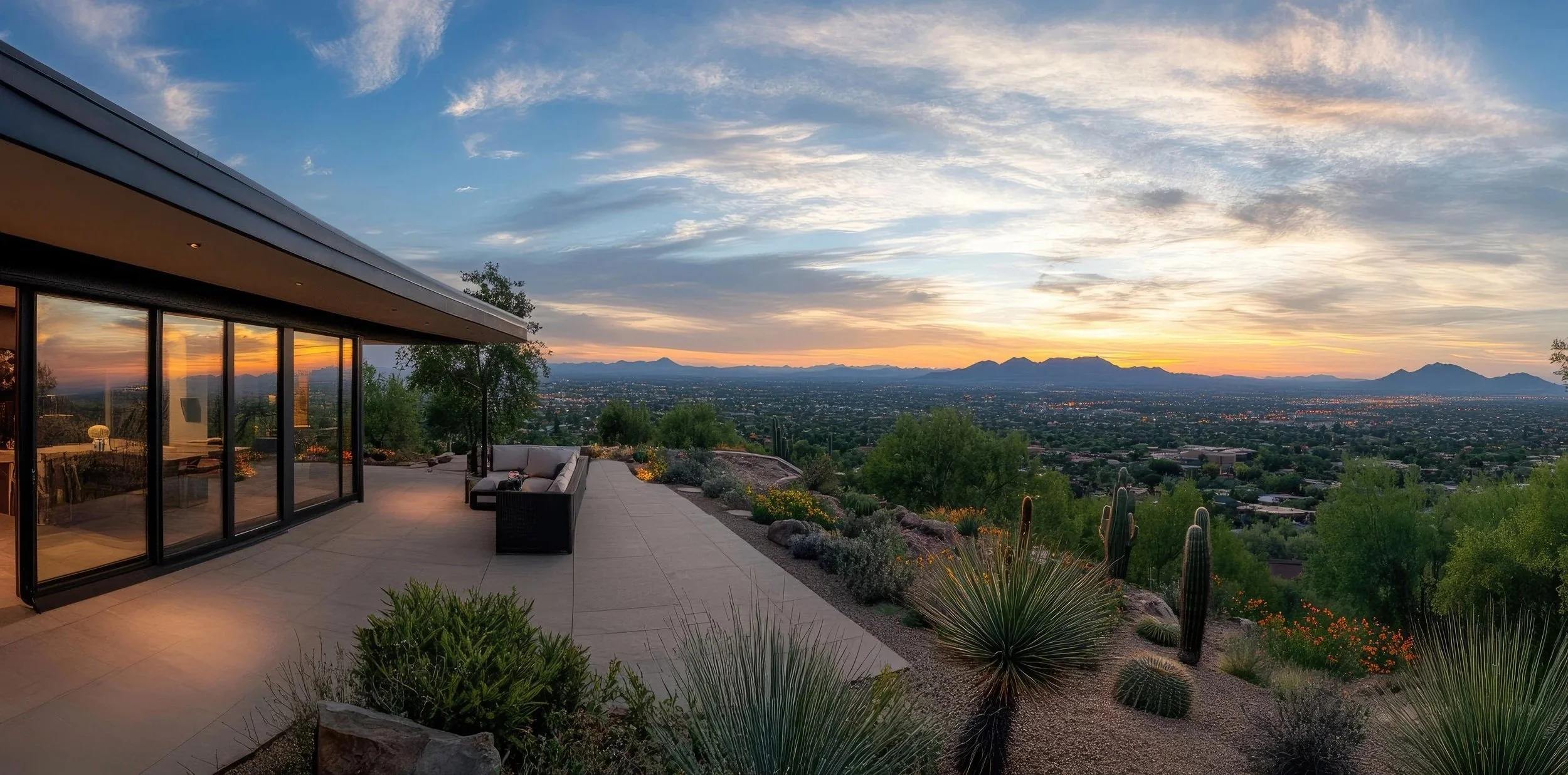 A scenic view of Scottsdale Arizona from a modern house patio at sunset, overlooking Phoenix with mountains in the background. The patio features sliding glass doors, outdoor seating, and desert plants.